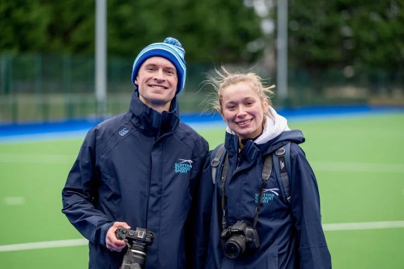 Two smiling young athletes in navy jackets and beanies, holding cameras, standing on a green sports field with a blue barrier and trees in the background.