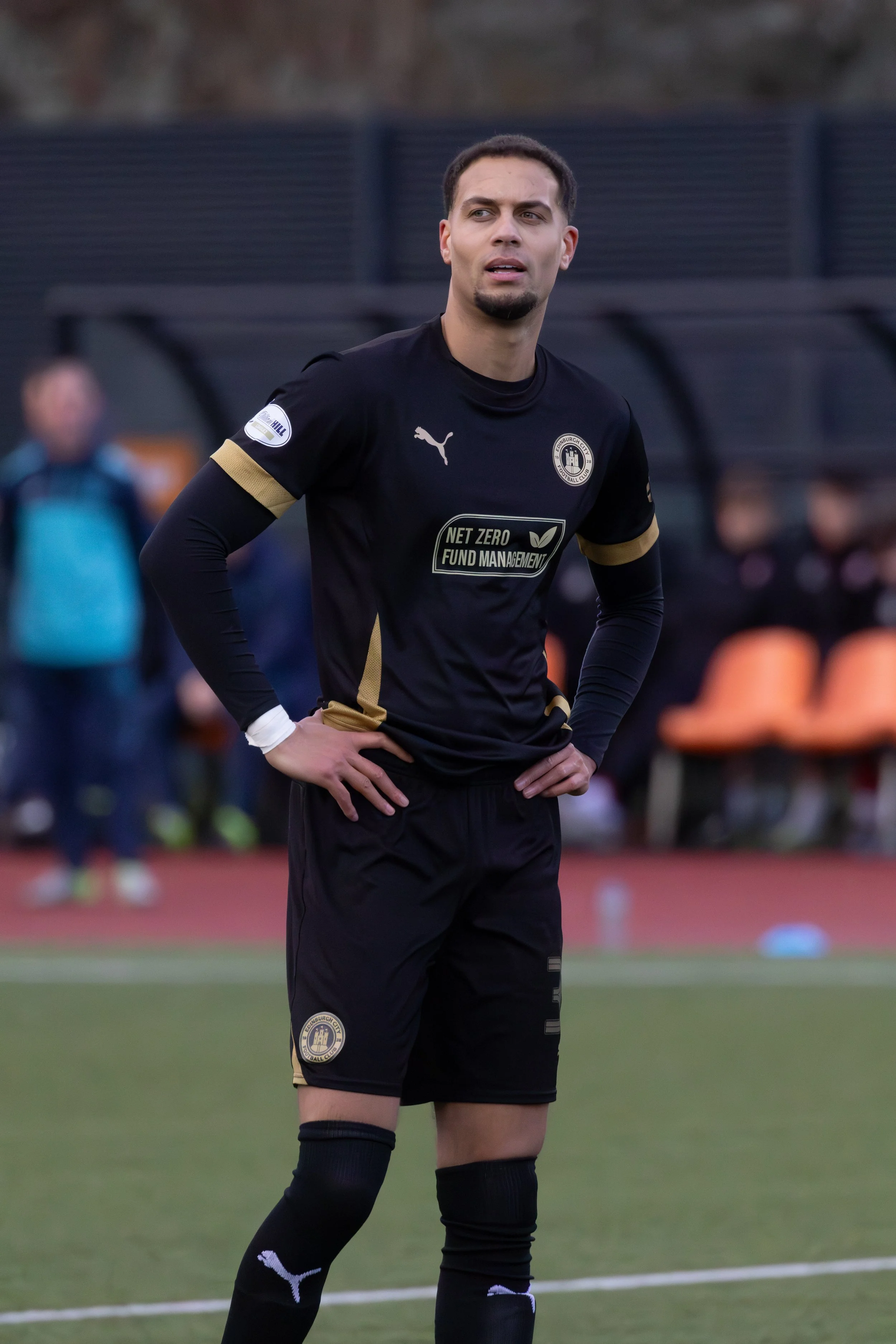 Soccer player standing on field with hands on hips, wearing black uniform with gold accents and sponsor logo.
