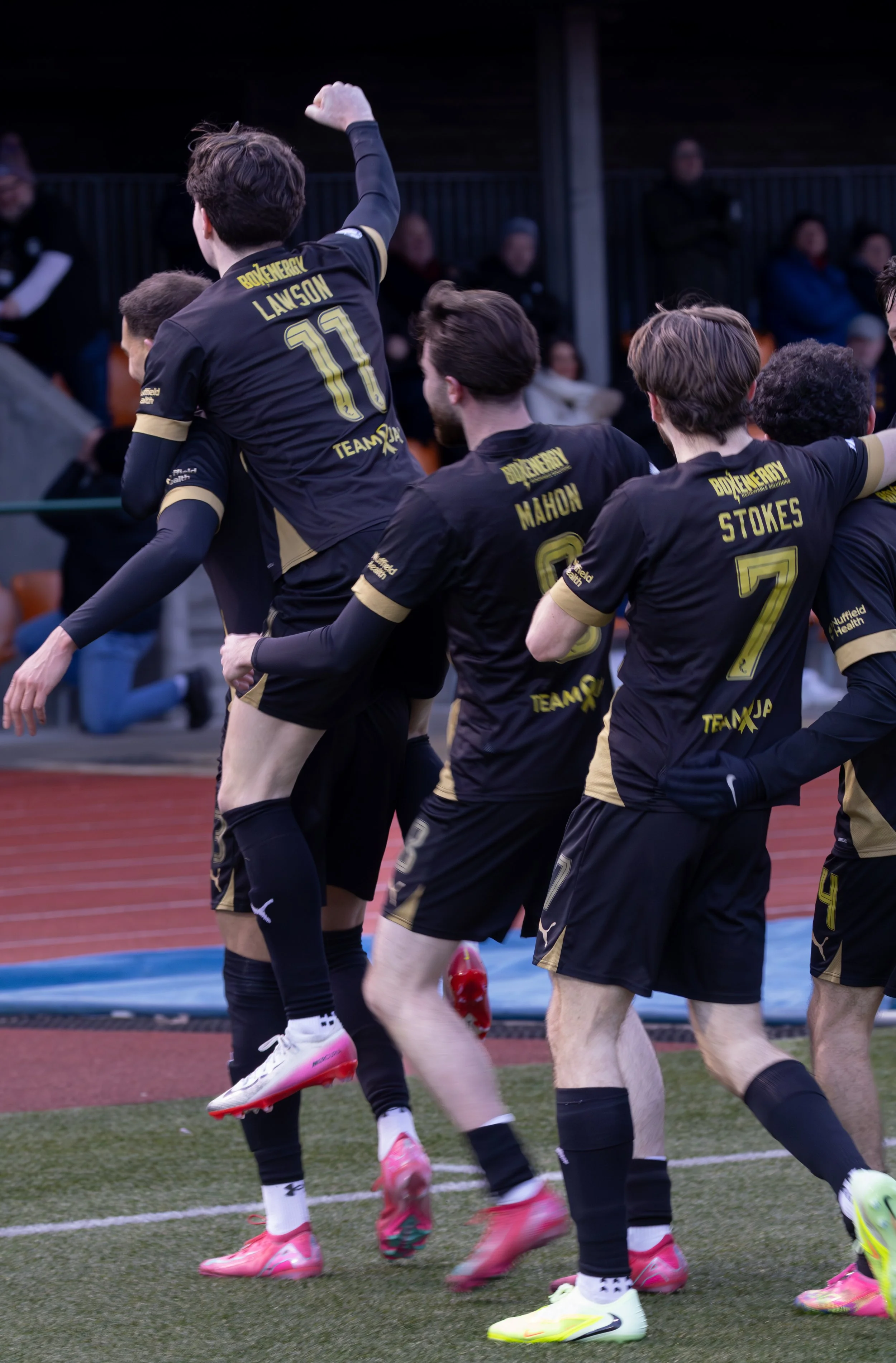 Soccer players in black and gold uniforms celebrating on the field, one player is being lifted by teammates.