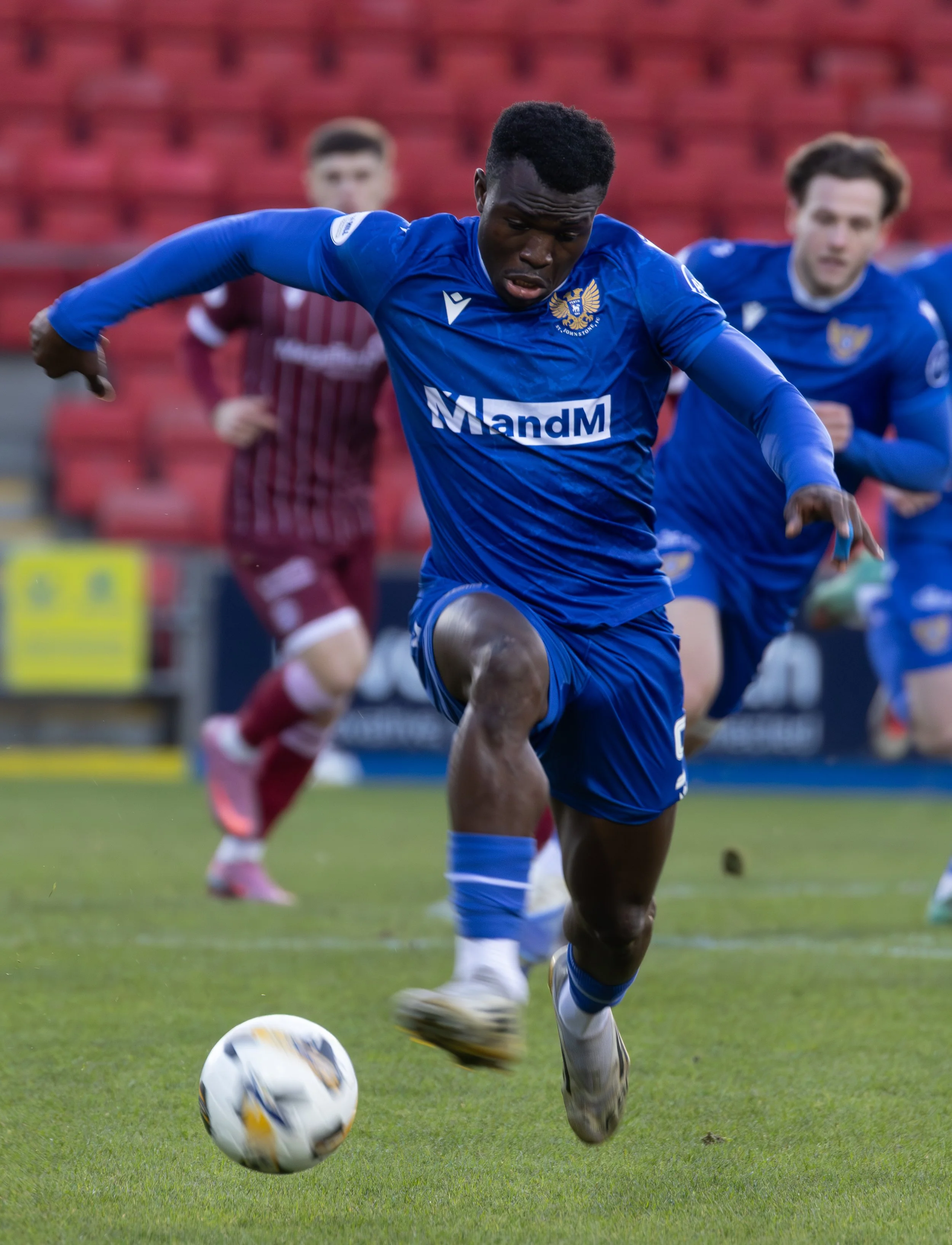 A soccer player in a blue uniform is running toward the ball on the field, with other players in the background.
