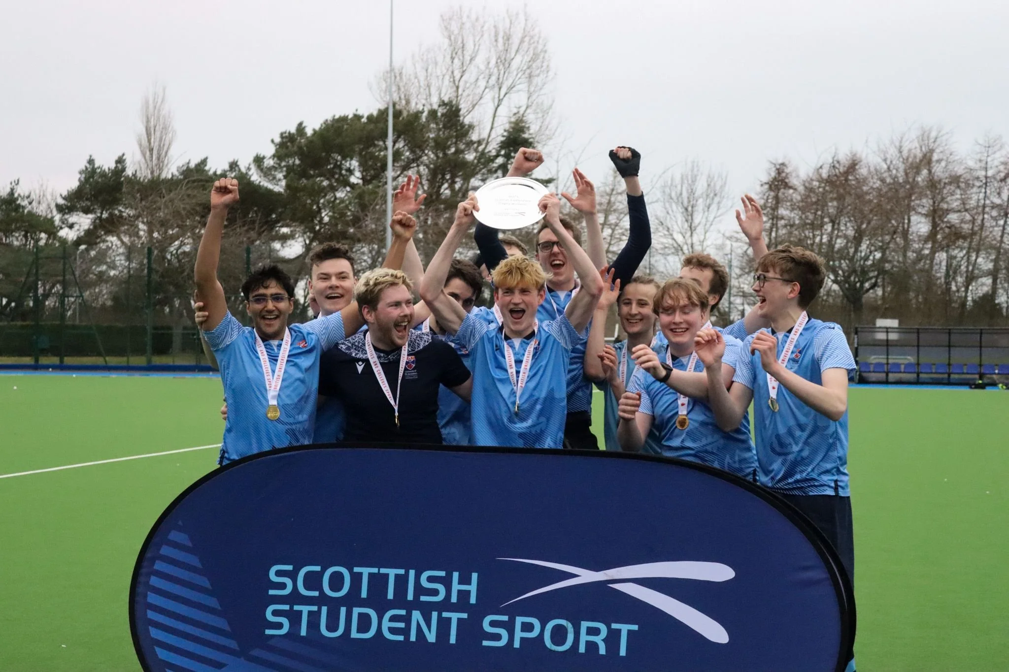 A group of young male athletes celebrating on a hockey field, holding a trophy and wearing medals, with a banner that reads 'Scottish Student Sport' in front of them.