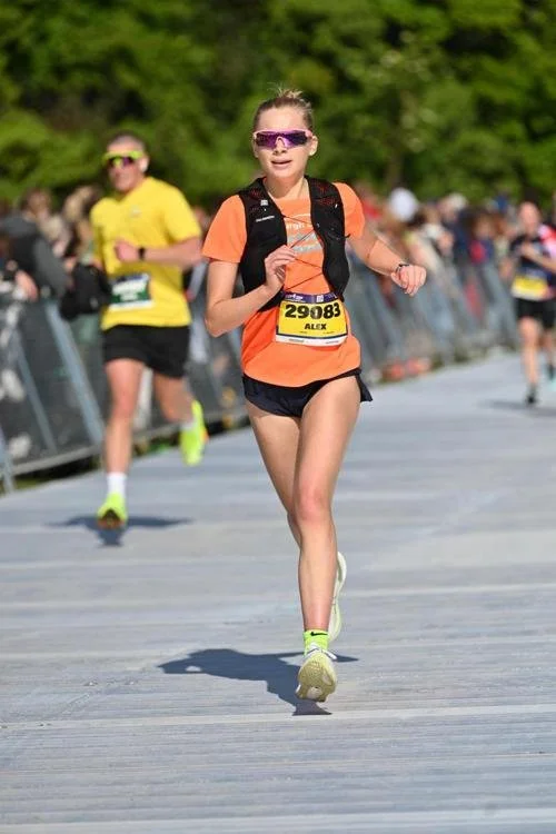 Young girl participating in a running race, wearing orange shirt, black shorts, purple sunglasses, and a black backpack, with a crowd and trees in the background.