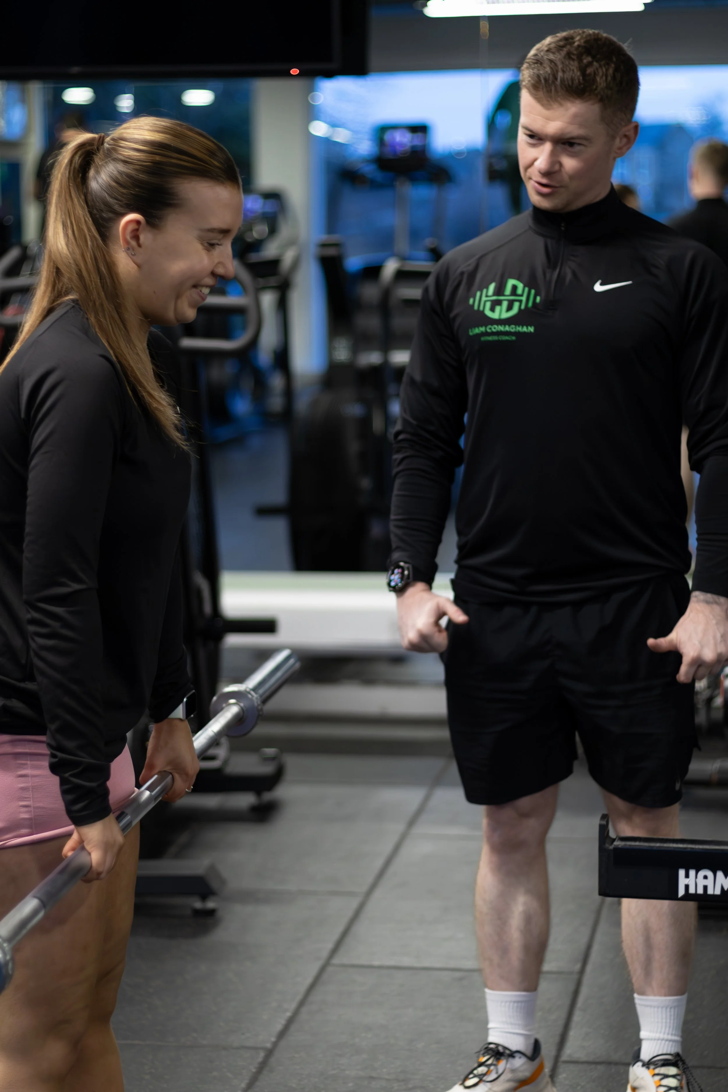 A woman and a man in a gym, the woman holding a barbell and smiling, and the man in athletic attire talking to her.