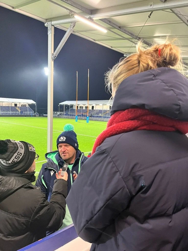 Three people are in a covered outdoor area at a sports stadium, with a football field visible in the background. A man in a navy jacket and beanie is giving a statement to two reporters holding microphones. The scene appears to be in the evening or u