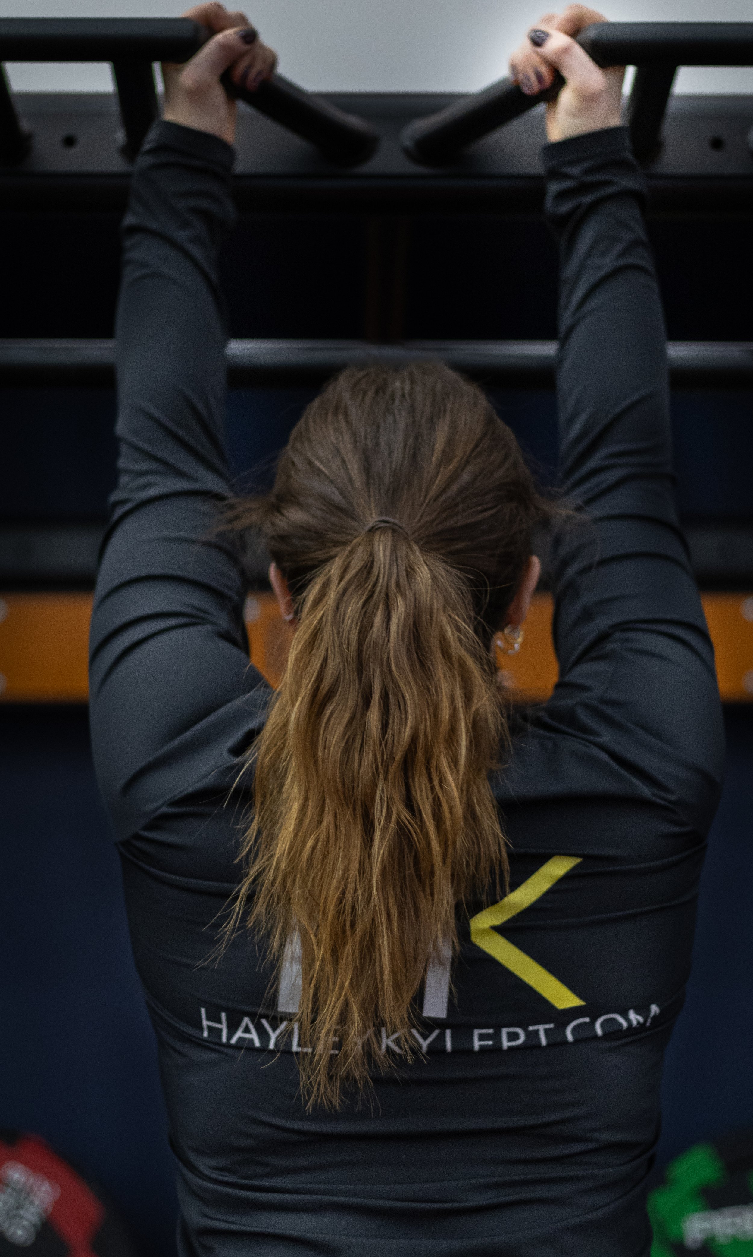 A woman with long brown hair tied in a ponytail wearing a black shirt and black pants, doing pull-ups on a bar in a gym.