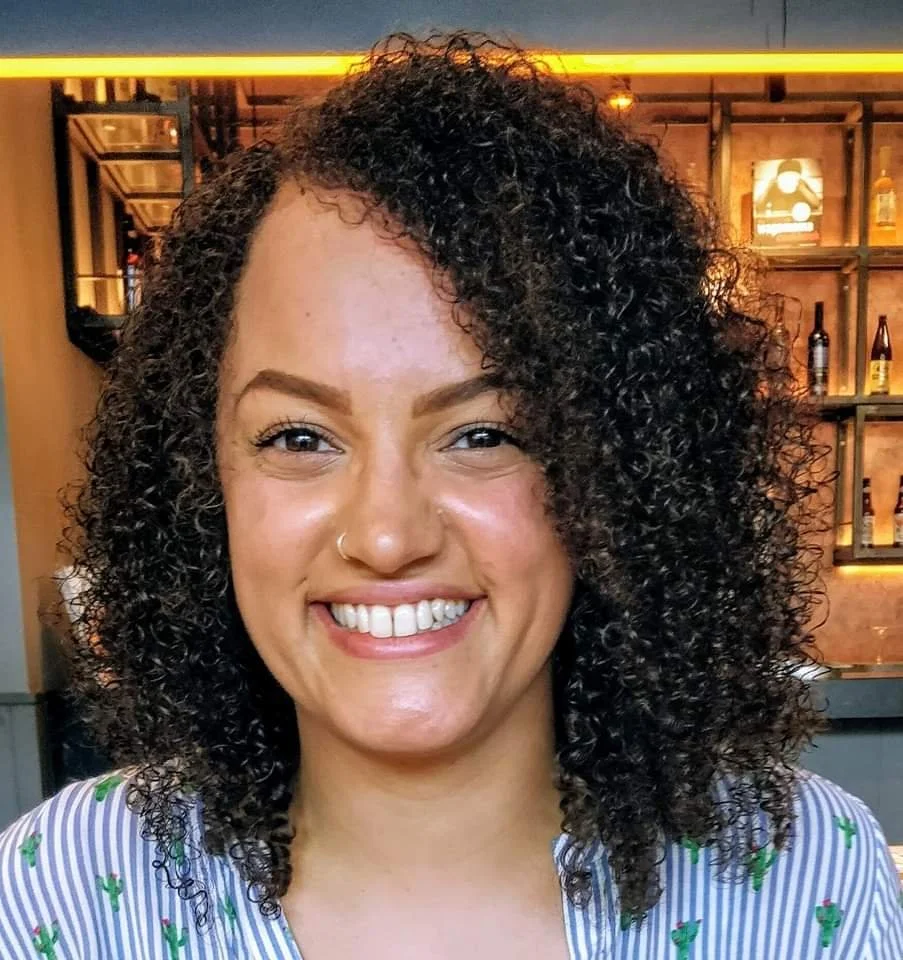 A smiling woman with curly dark hair, wearing a striped shirt with small green cactus patterns, is in a bar or restaurant with bottles on shelves in the background.