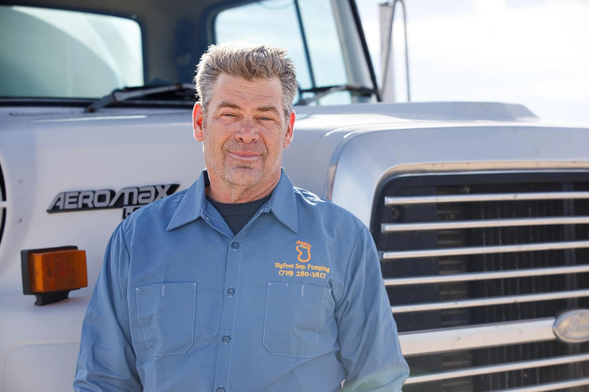 A man in a blue work shirt standing in front of a large white truck with the logo "AEROMAX" on it. The shirt has a logo with a footprint and the text "BigFoot Sept. Pumping" along with a phone number.