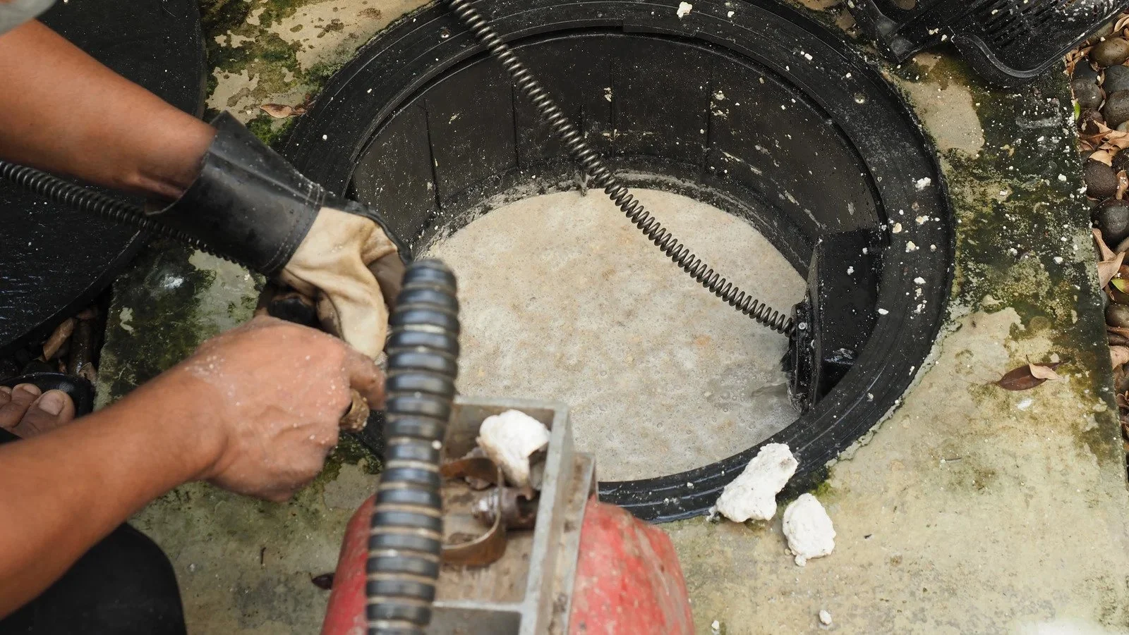 Person wearing gloves using a tool to clear out a clogged drain or pipe in a black circular outdoor drain with water and debris around it.