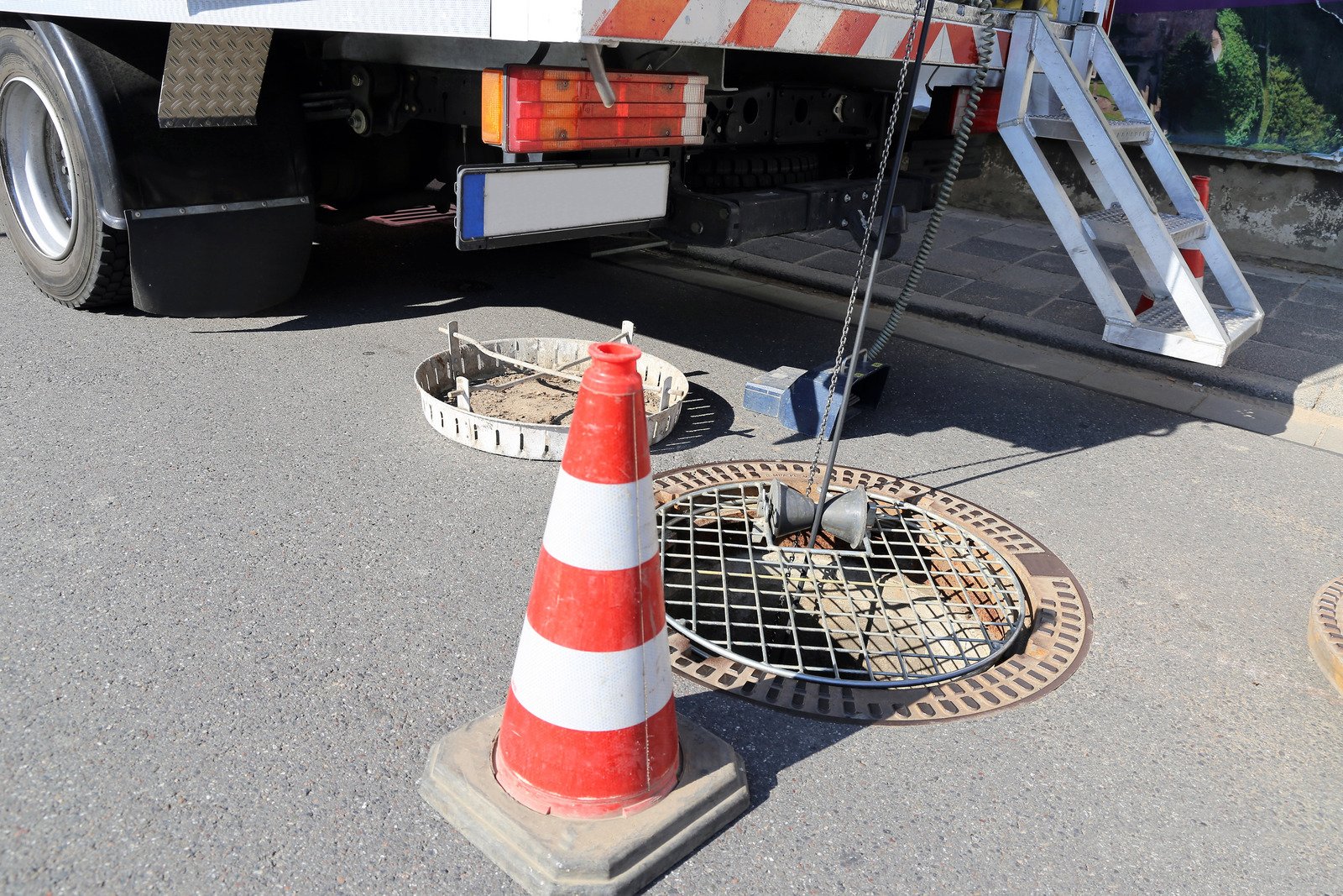 Construction or utility work scene showing a street with a traffic cone, a manhole, a caution sign, traffic camera, and a truck with a lift ladder.