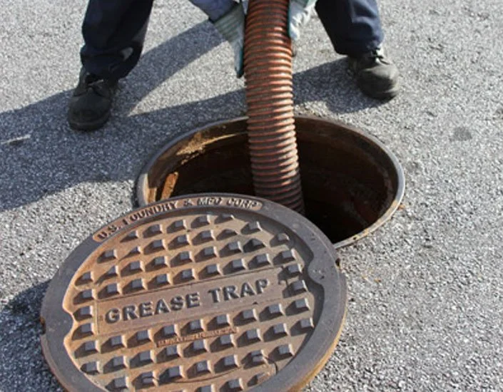 A person standing over an open manhole with a flexible hose coming out of it. The manhole cover, which is on the ground beside its opening, is labeled 'GREASE TRAP' and has a grid pattern.