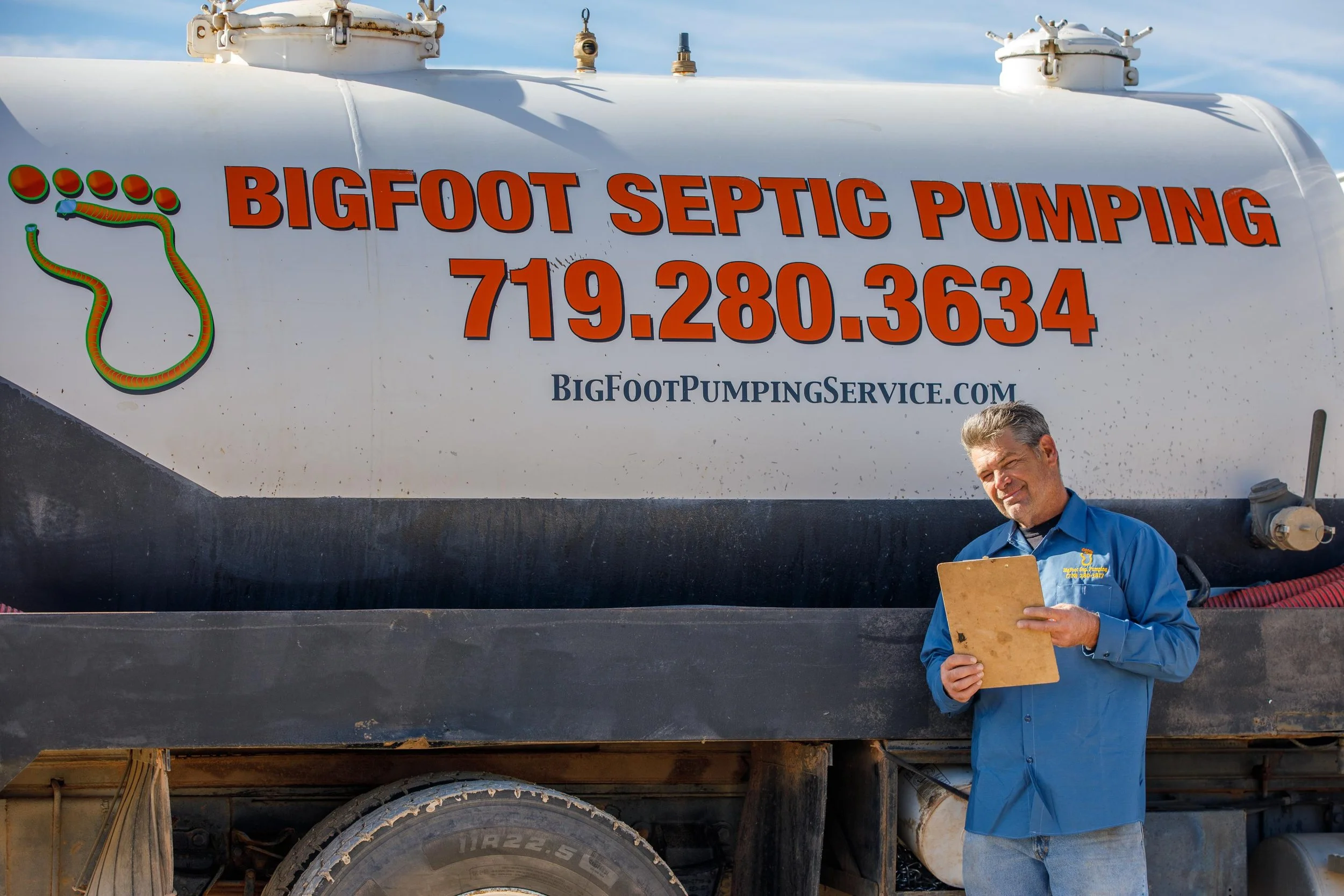 A man standing next to a large tank labeled Bigfoot Septic Pumping, holding a clipboard and looking at it, with the contact number and website for the septic service on the tank.