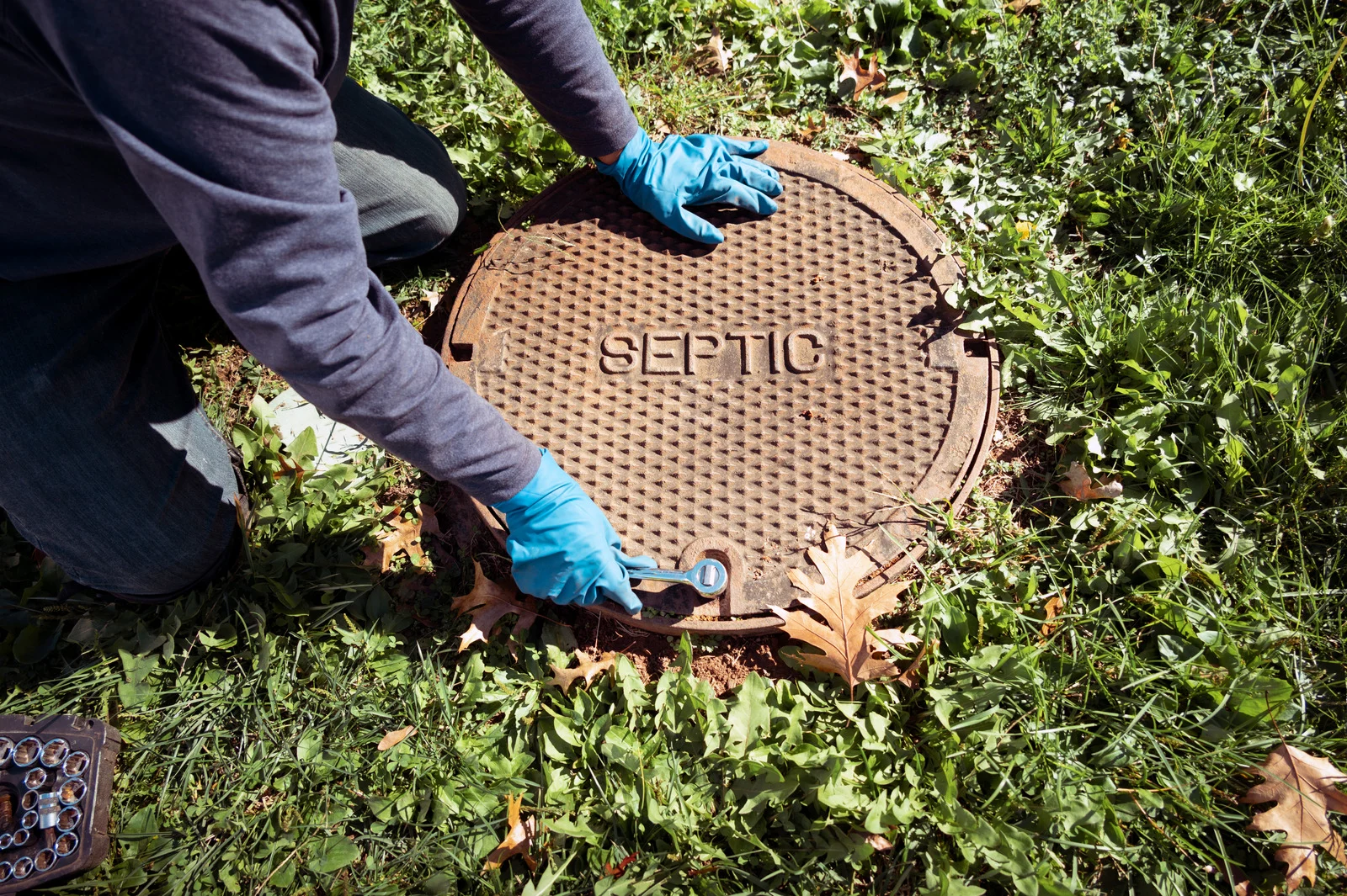 A person wearing blue gloves is using a wrench to open a septic tank cover labeled 'SEPTIC' surrounded by grass and fallen leaves.