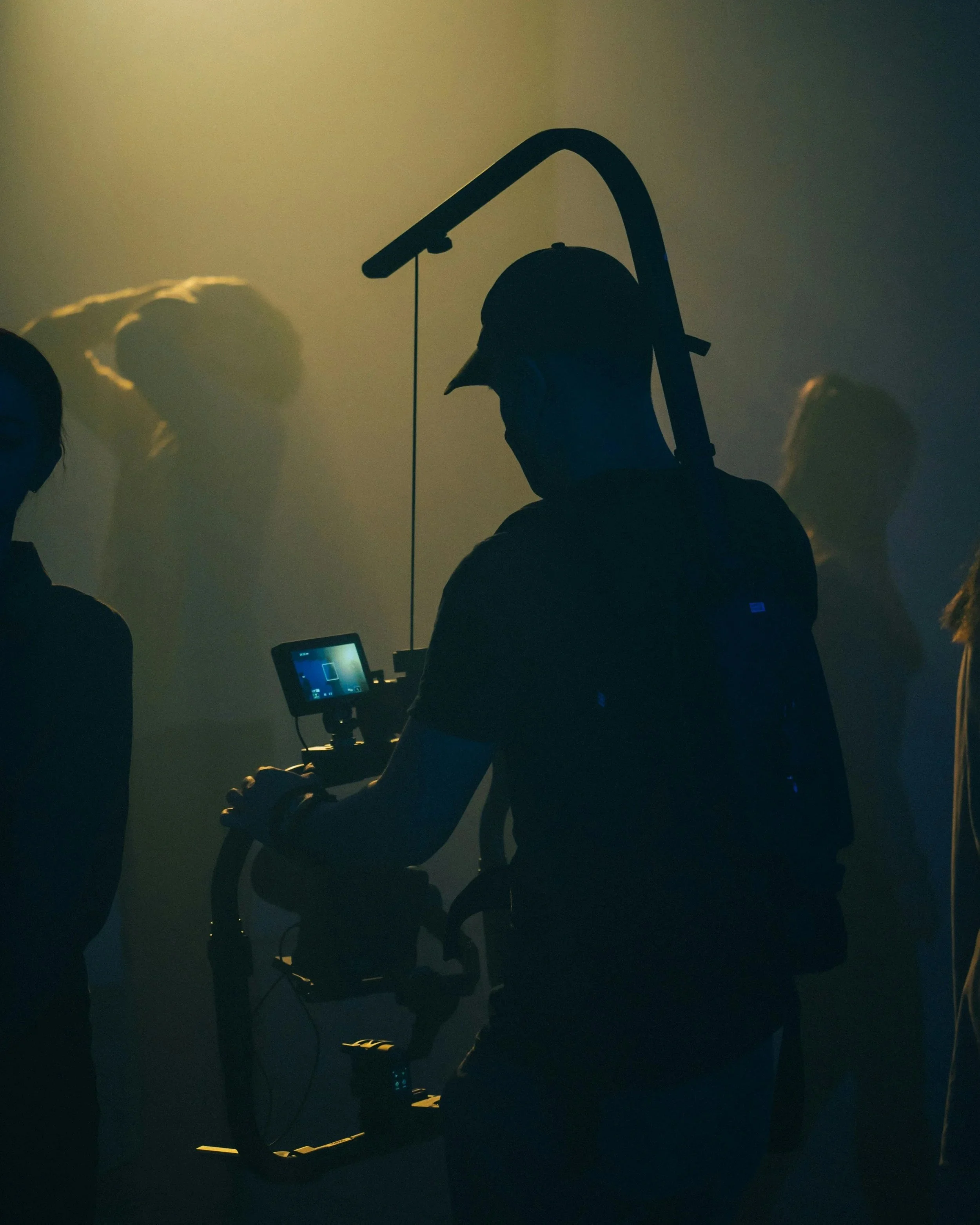 Silhouette of a person with a helmet and harness operating a camera in a dimly lit room with other people in the background.