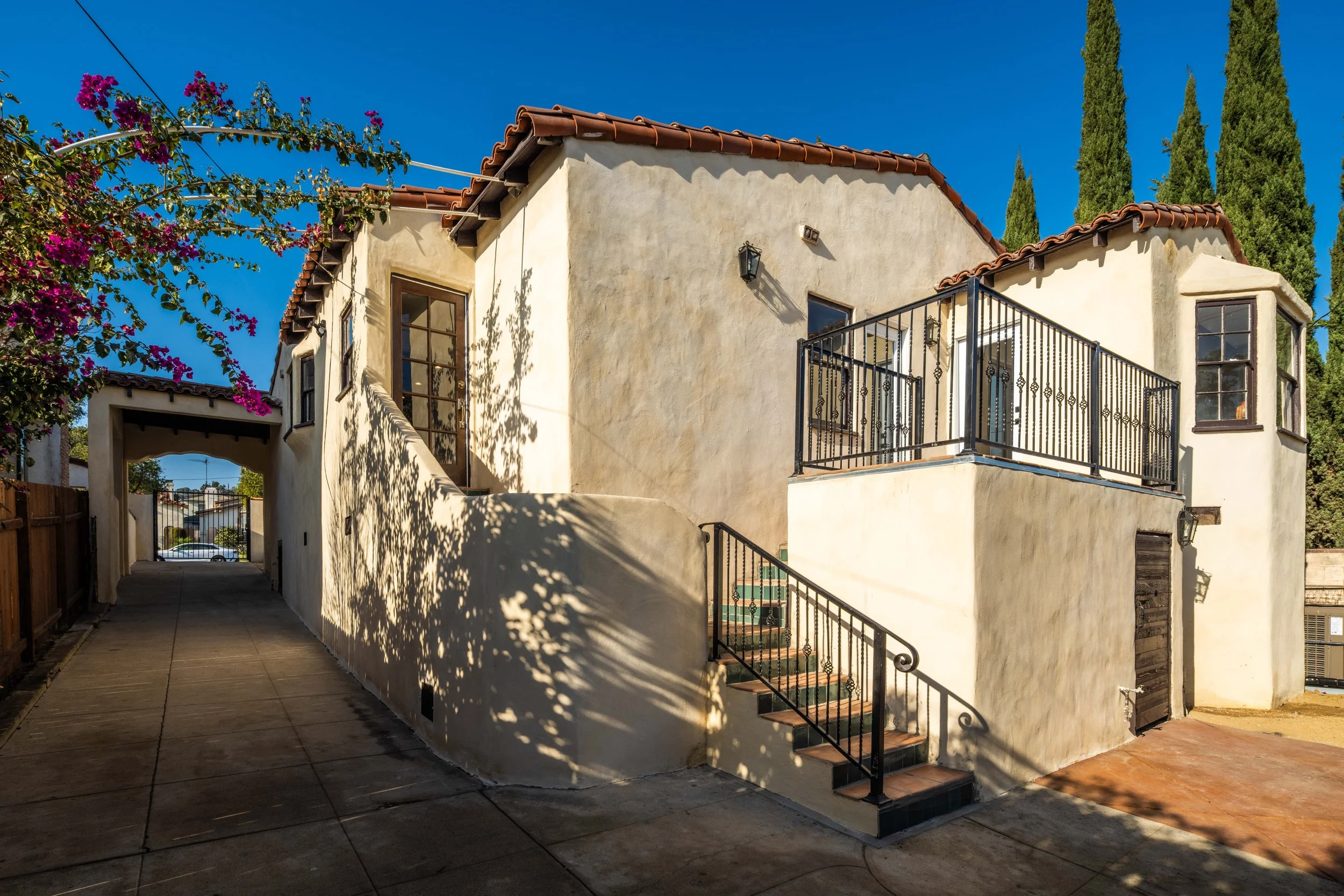 A Mediterranean-style beige stucco house with red tiled roof, iron balcony, and stairs leading to a side door, surrounded by trees and a clear blue sky.