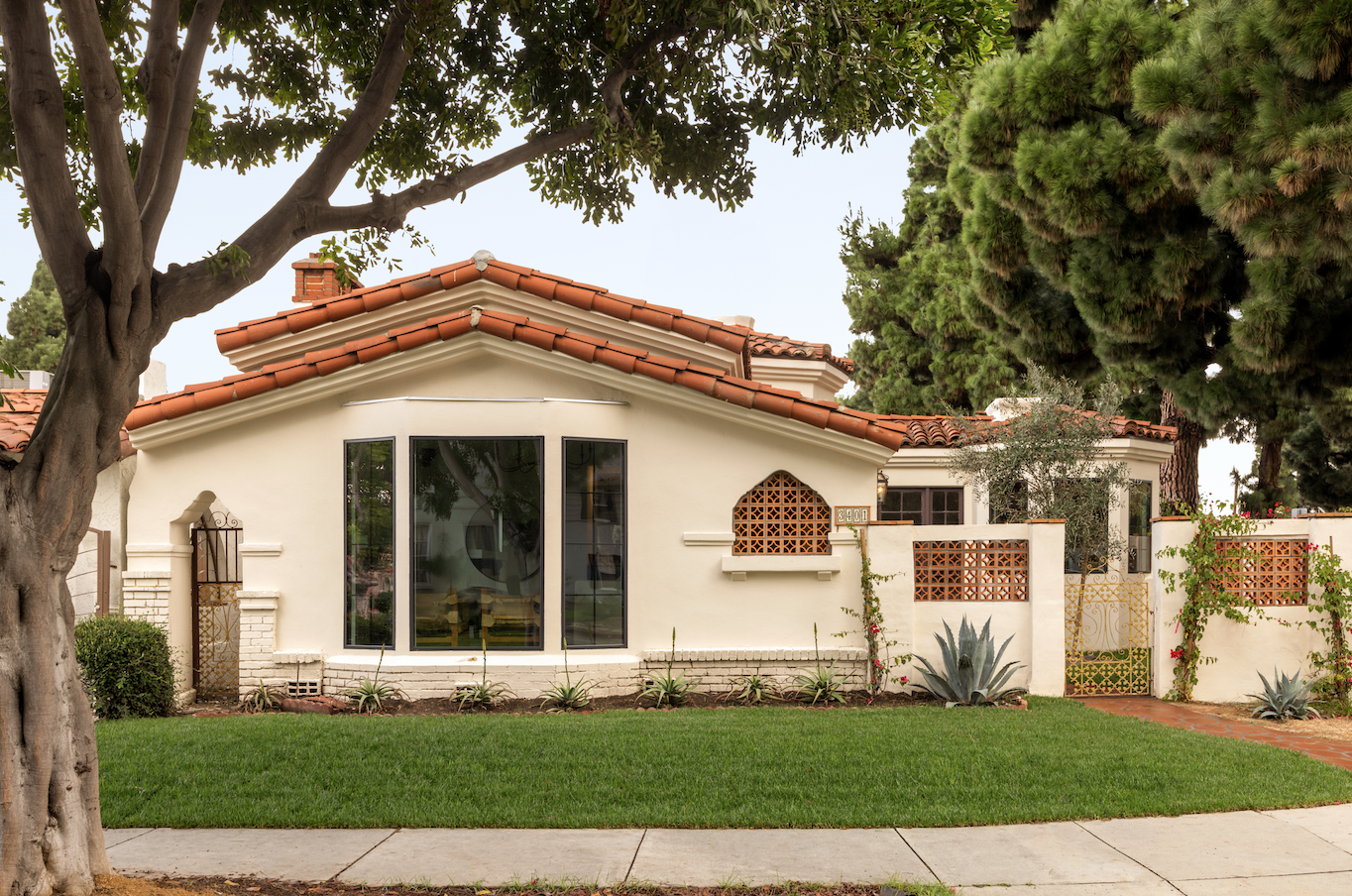 Front yard of a house with a manicured lawn, large trees, and a white stucco exterior with red tile roof and decorative brickwork.