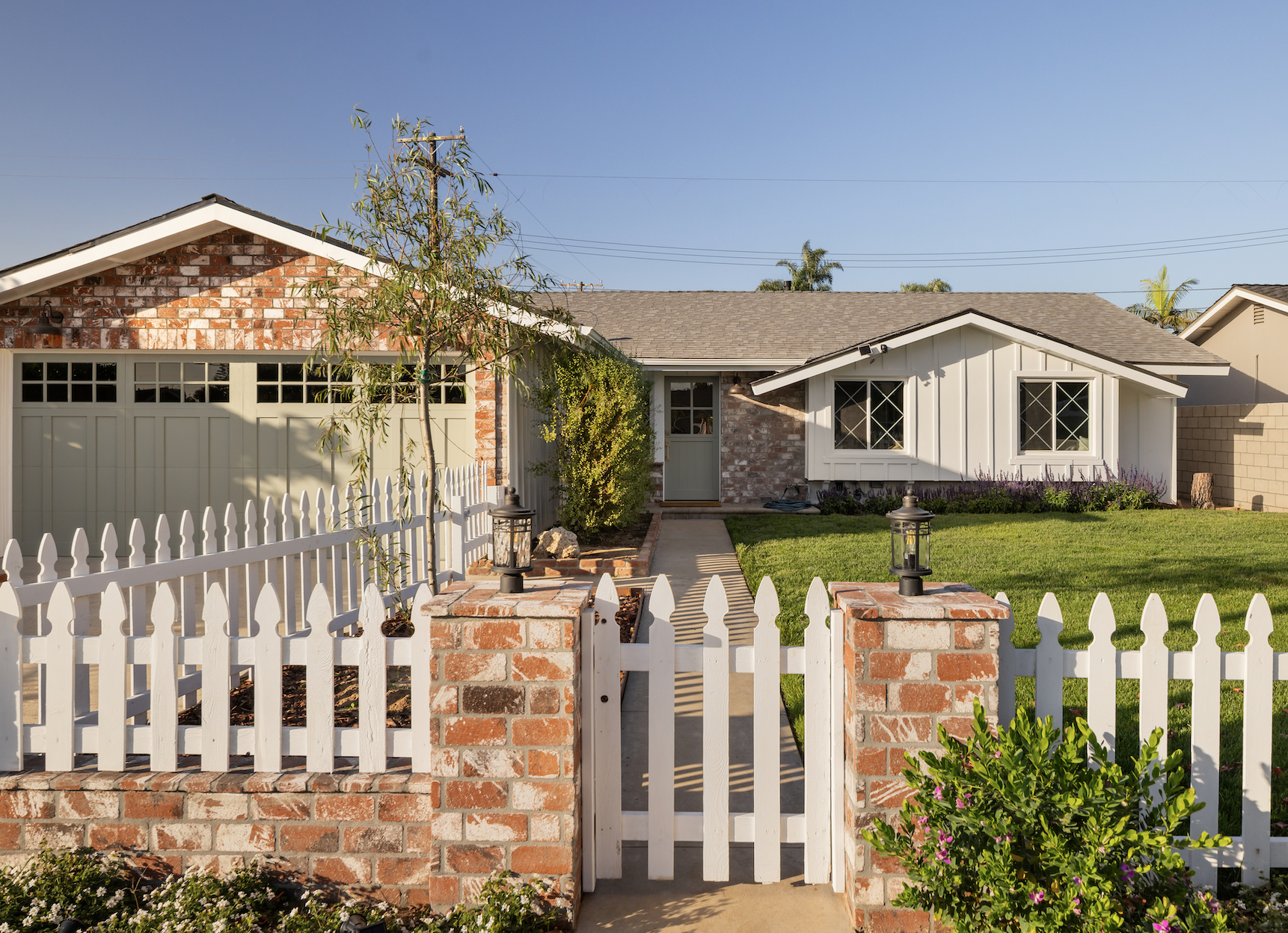 A house with a white picket fence, brick columns, a green lawn, a concrete walkway, and a detached garage, with a clear sky in the background.