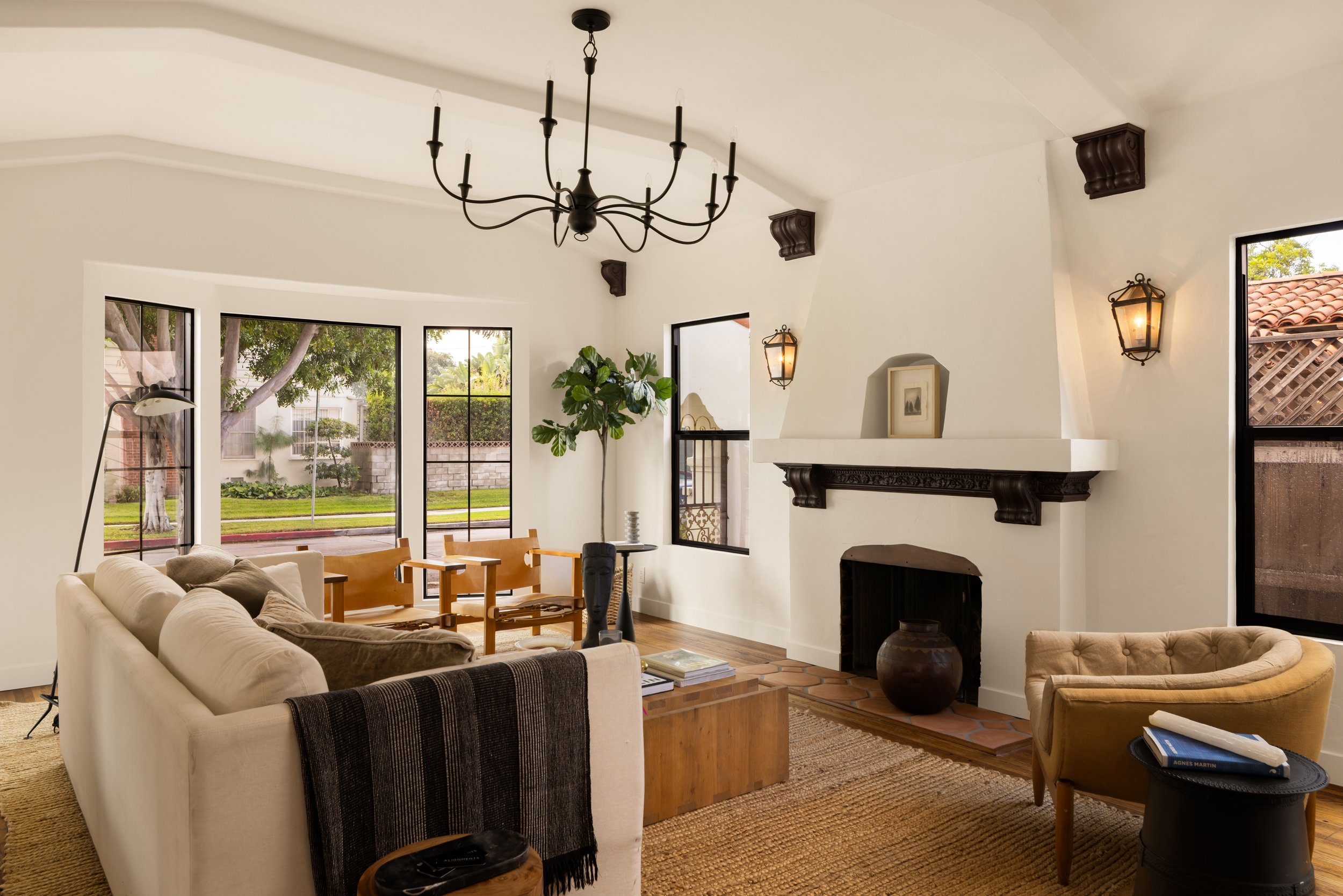 Living room with white walls, large windows, a beige sofa, a yellow armchair, a black fireplace, a black chandelier, wall sconces, a green potted plant, and a wooden floor.