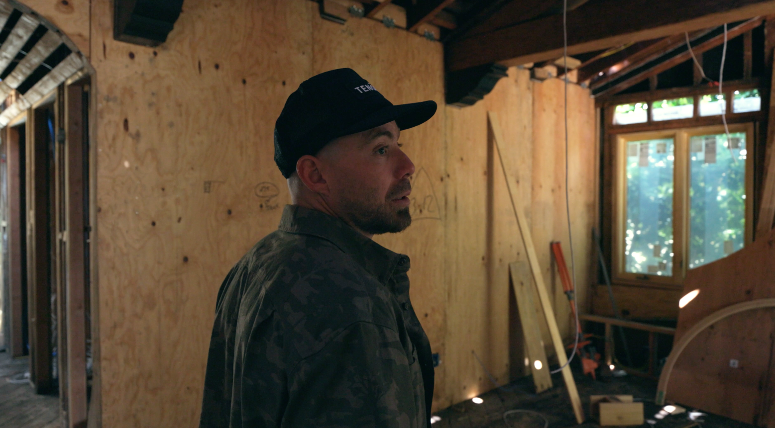 Man in a black cap and camouflage jacket inside a construction or renovation site with plywood walls, windows, and tools.