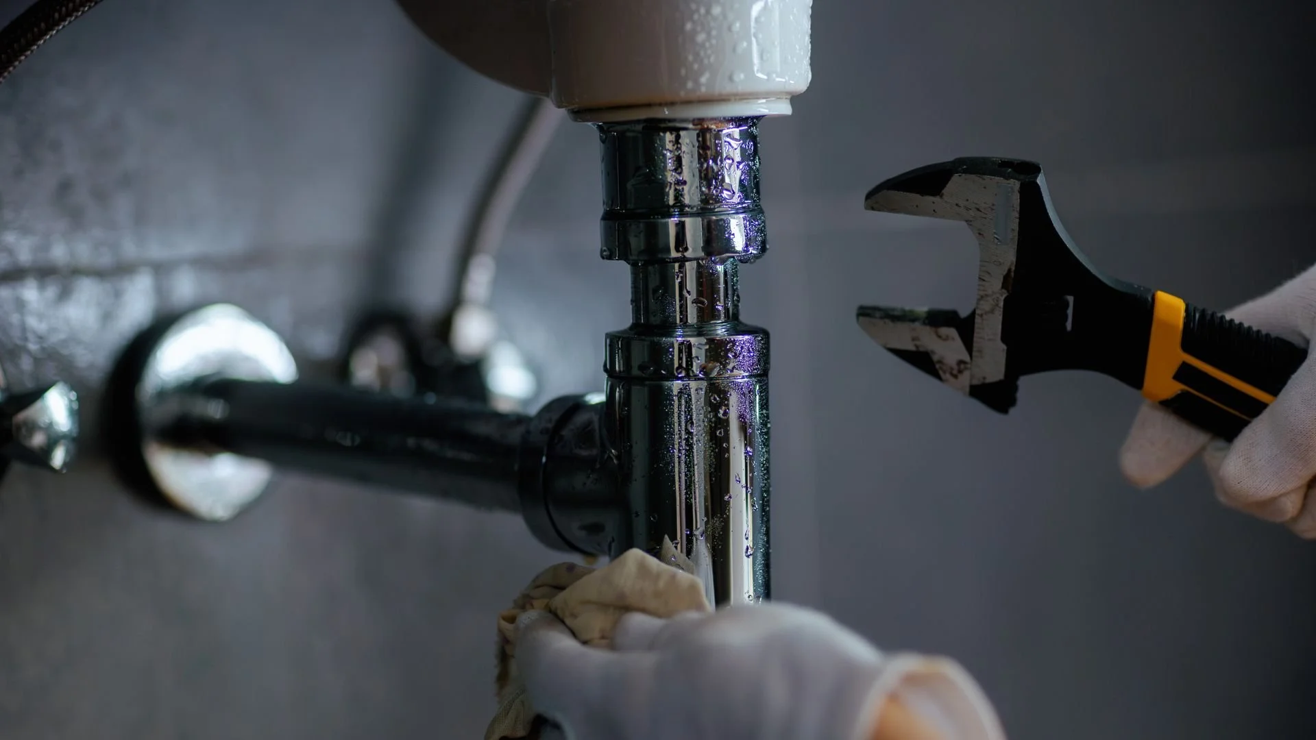 A person wearing gloves uses a wrench to fix plumbing pipes under a sink.