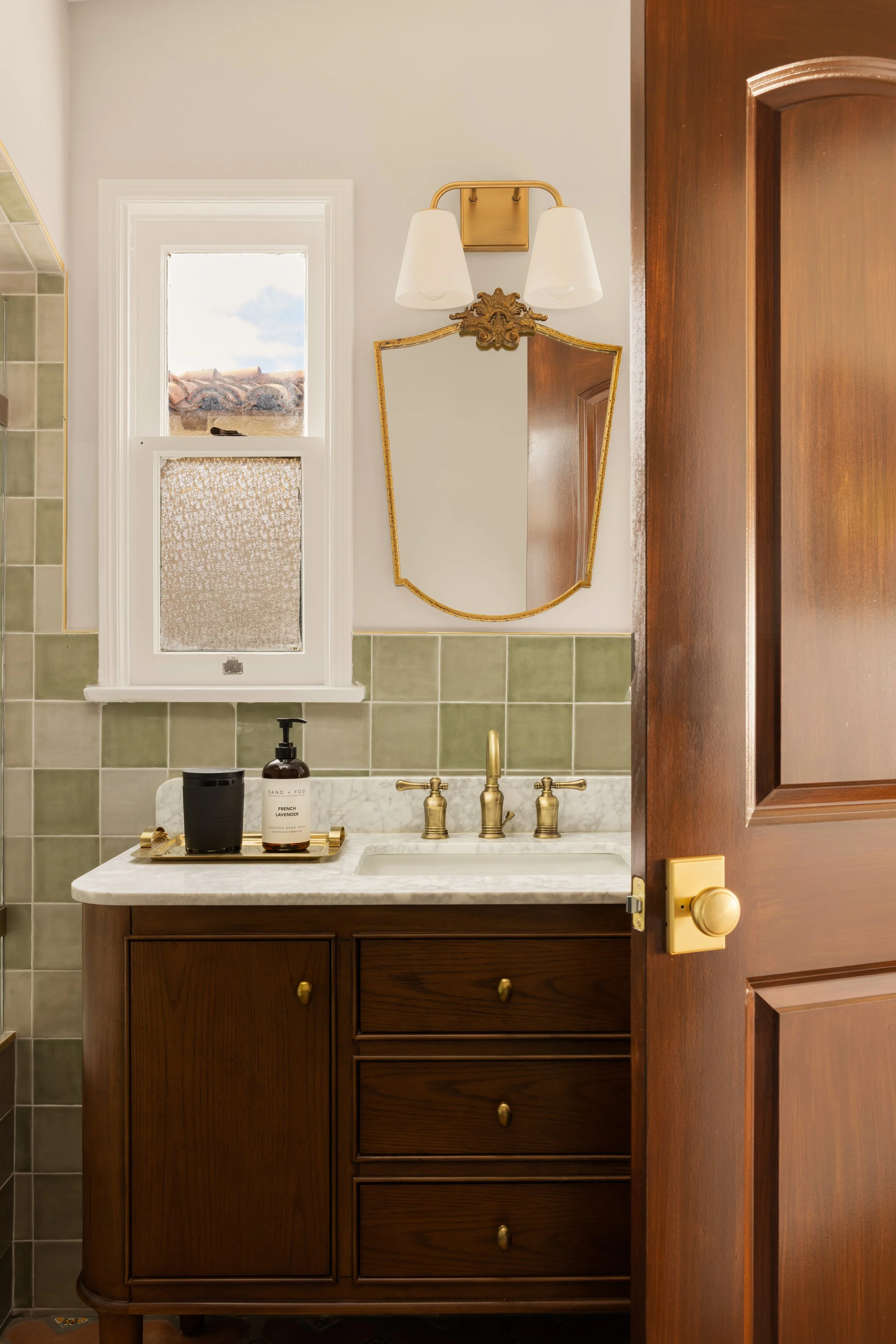 A vintage-style bathroom vanity with a white marble top, brass fixtures, and a small mirror. The vanity has dark wood cabinets and drawers. There is a small window above the sink, a candle and a bottle of soap on a tray, and a wall-mounted light with two white lampshades.