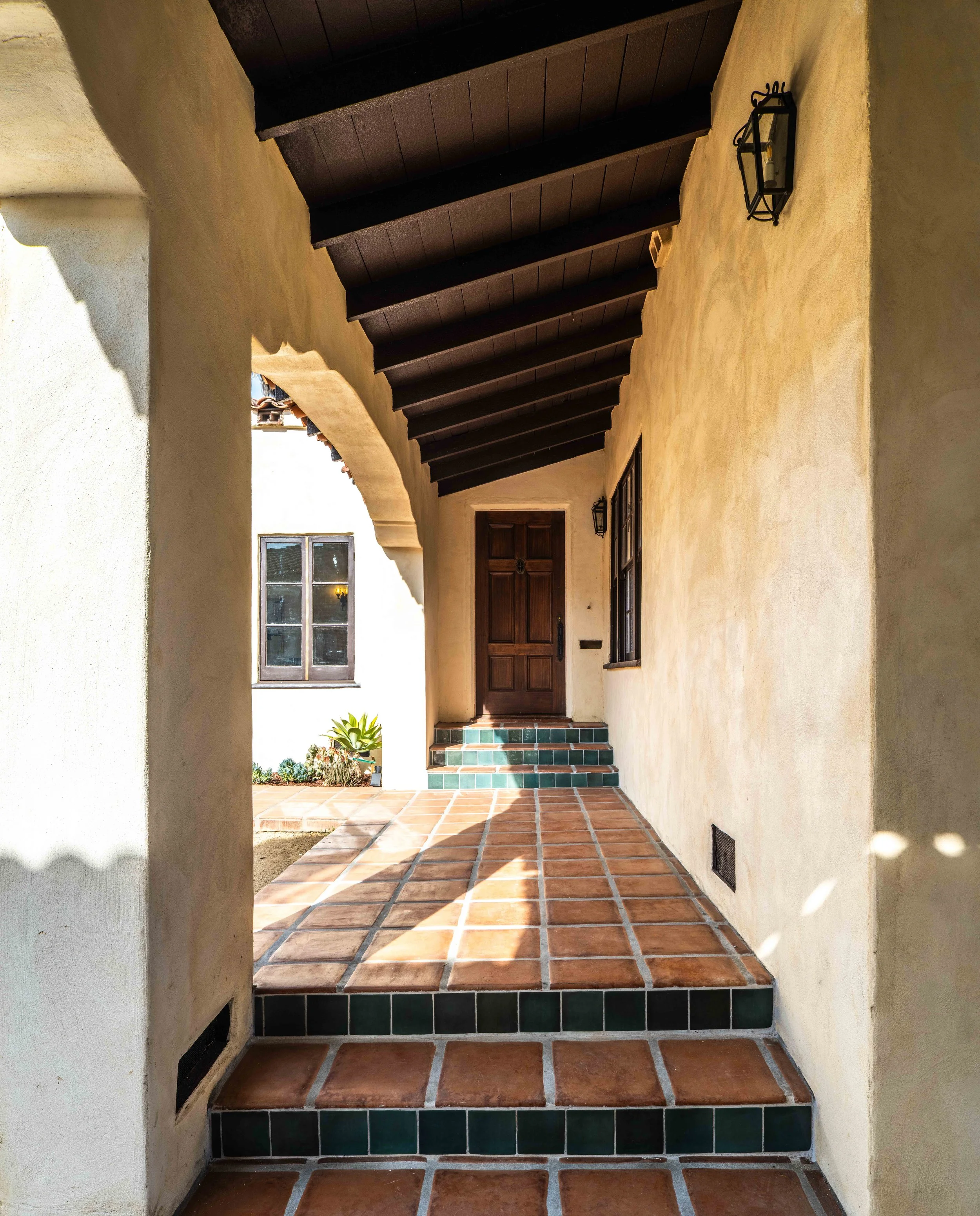 View of a Spanish-style porch with terracotta tile flooring, arched entryway, wooden ceiling beams, a dark wooden front door, and black wall-mounted lanterns.