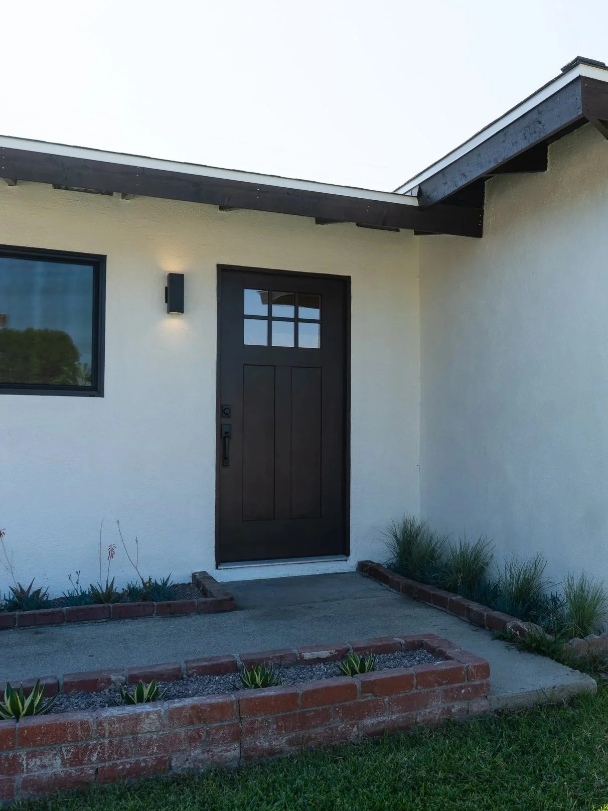 Front entry door of a modern house with a black door, small window above, exterior wall light, and a simple garden bed with small plants and bricks in front.