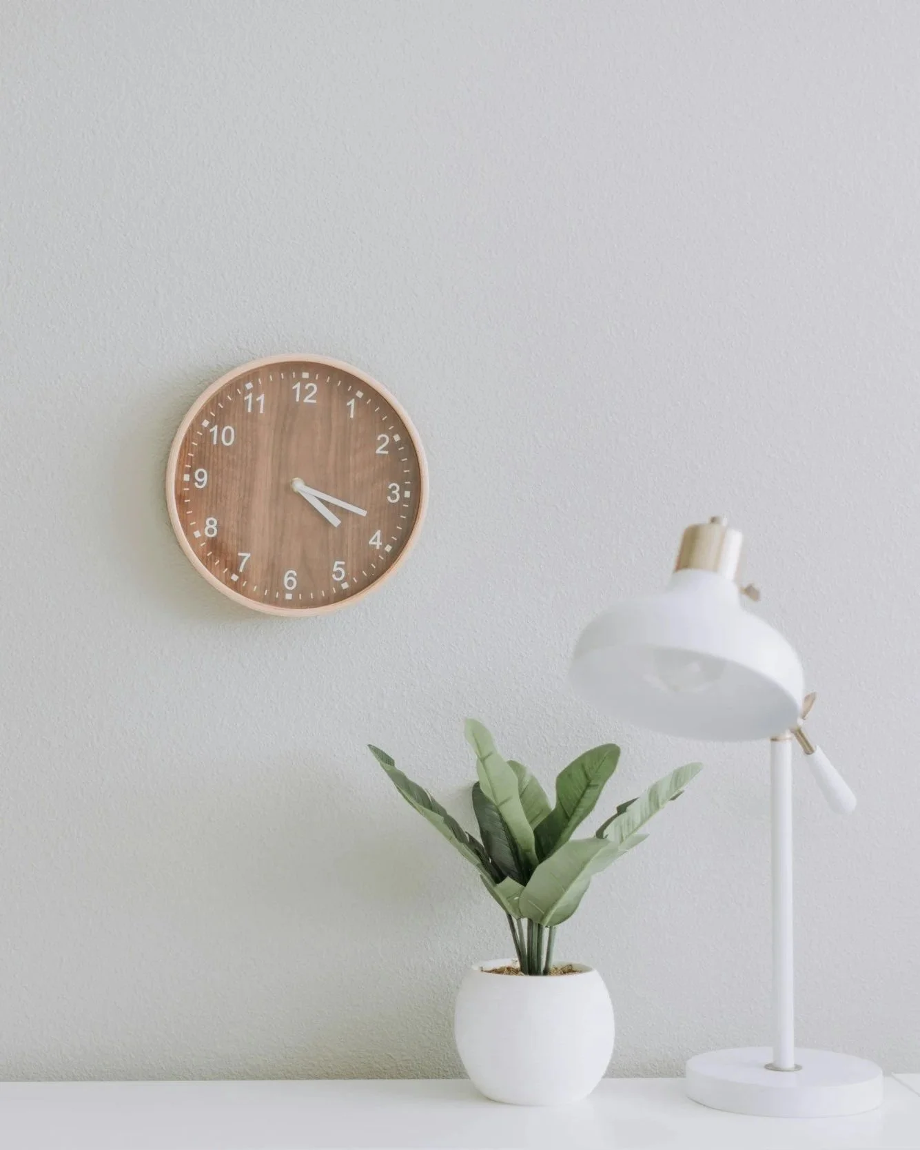 A round wall clock with a wood finish face showing approximately 3:20, a white pot with green leafy plant, and a white desk lamp on a white surface against a light-colored wall.