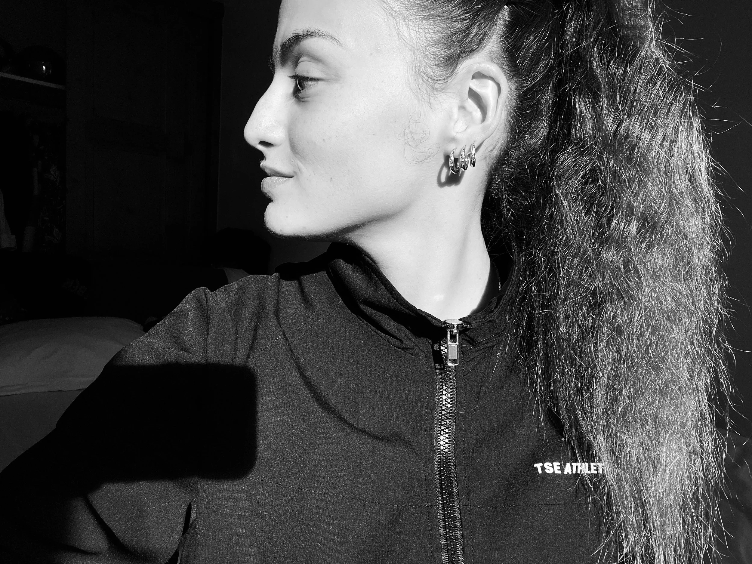Portrait en noir et blanc d'une femme avec de longs cheveux ondulés, portant un sweat à fermeture éclair avec le logo 'TSE ATHLETICS', regardant vers la gauche.