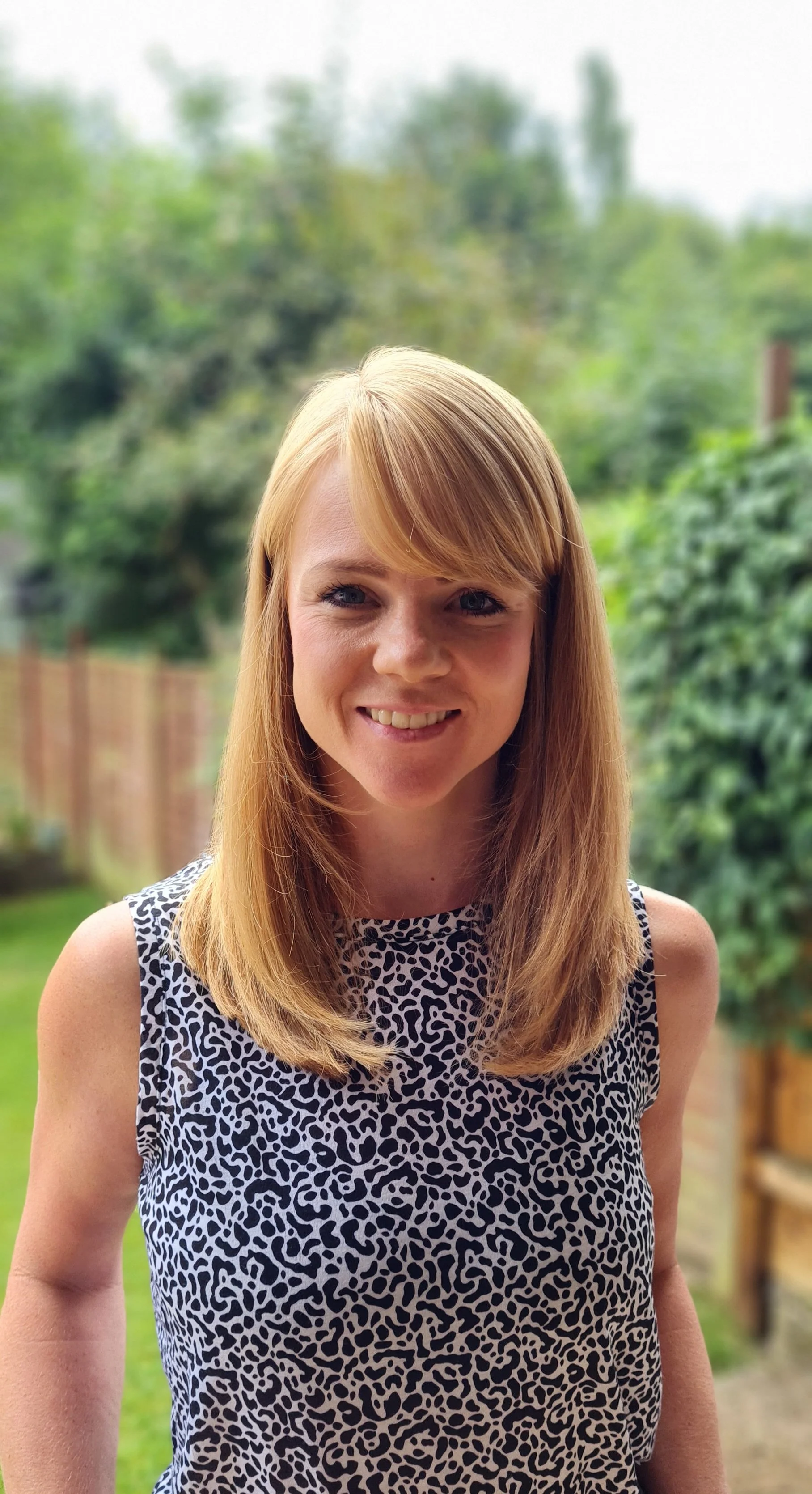 A woman with long blonde hair smiling outdoors, wearing a sleeveless black and white patterned top, with a blurred green garden background.