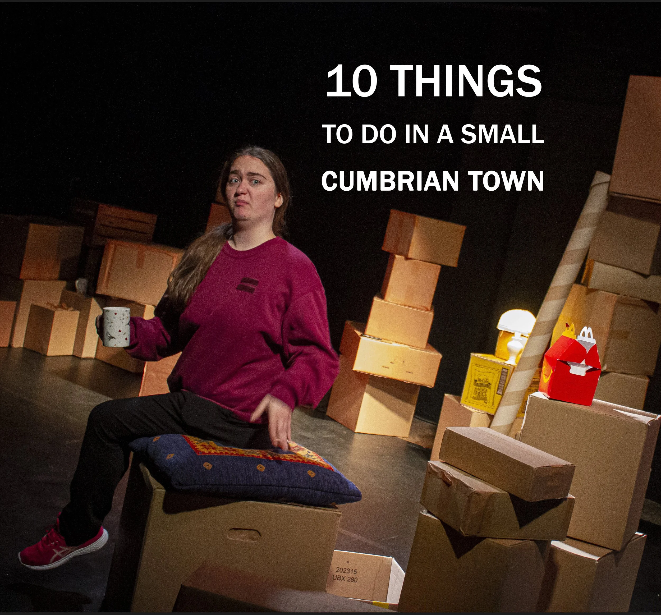Scene from a theatrical performance with a woman holding a mug, sitting on a box with a pillow, surrounded by cardboard boxes and props, with text overlay reading '10 Things To Do in a Small Cumbrian Town.'