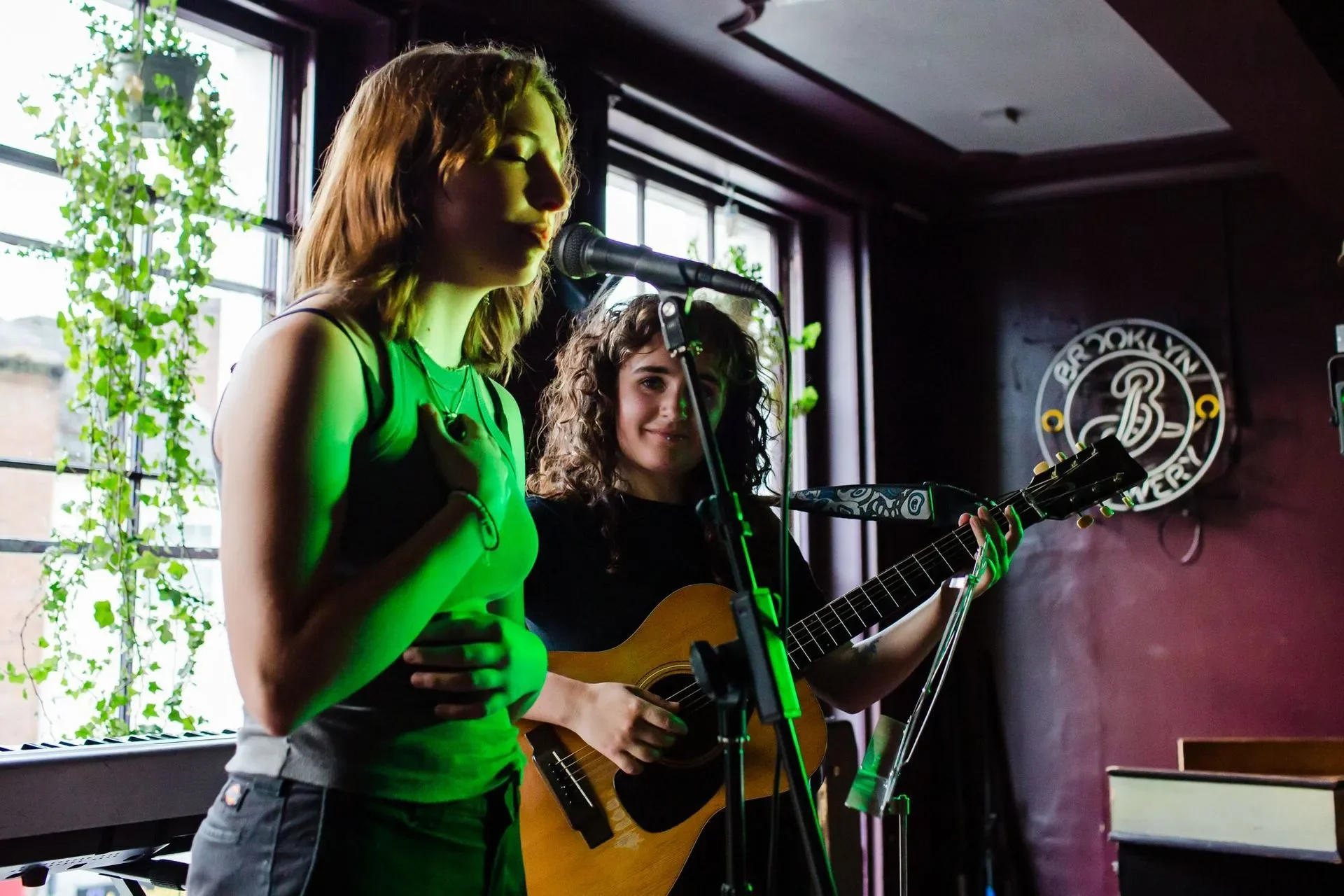 Two young women performing music in a dimly lit venue, one singing with her hand on her chest and the other playing guitar, with a window and greenery in the background.