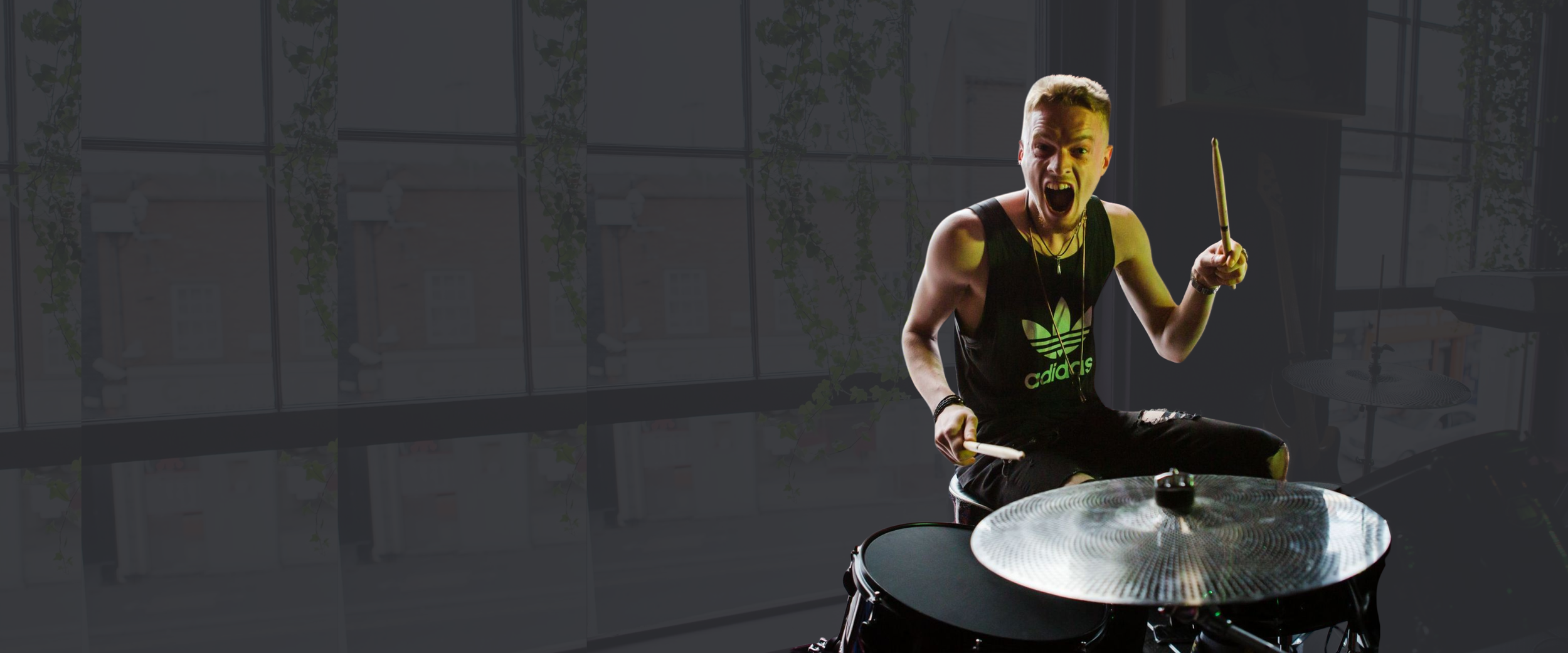 A young man with short hair and a sleeveless black Adidas shirt is playing drums indoors, expressing excitement with his mouth open and holding drumsticks.