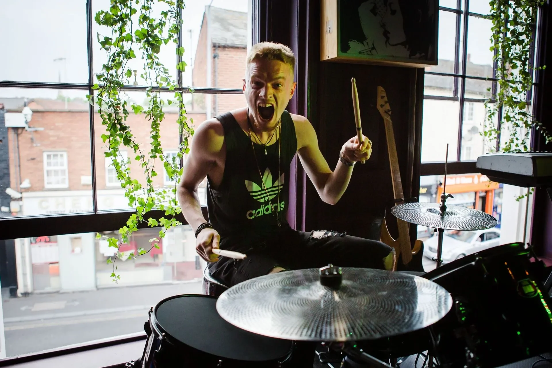 Young man with bleached hair playing drums enthusiastically indoors near a large window, with plants and guitars in the background.