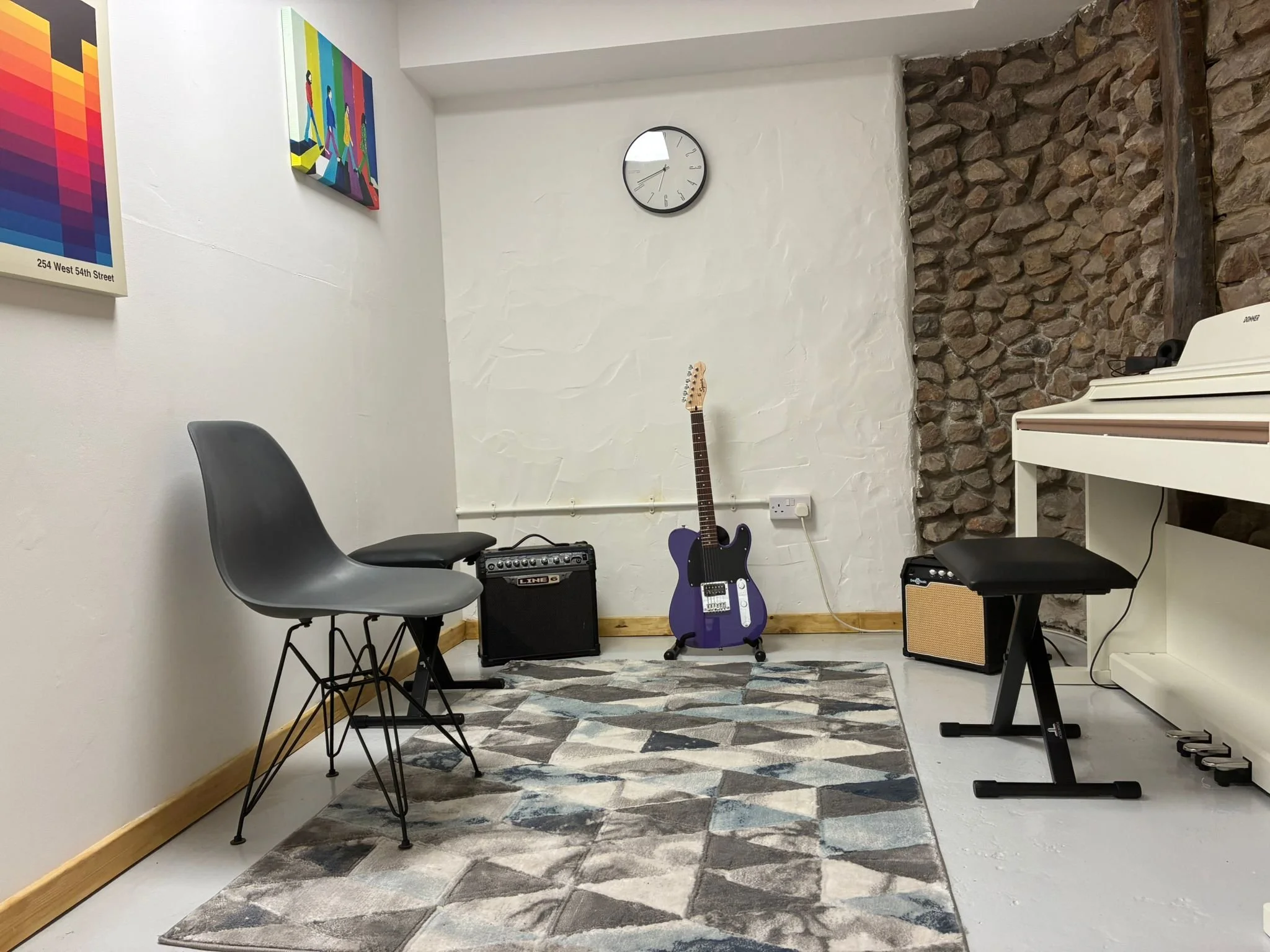 Music room with a purple electric guitar, two small amplifiers, a piano, a stool, a modern chair, a clock, and colorful artwork on the walls.