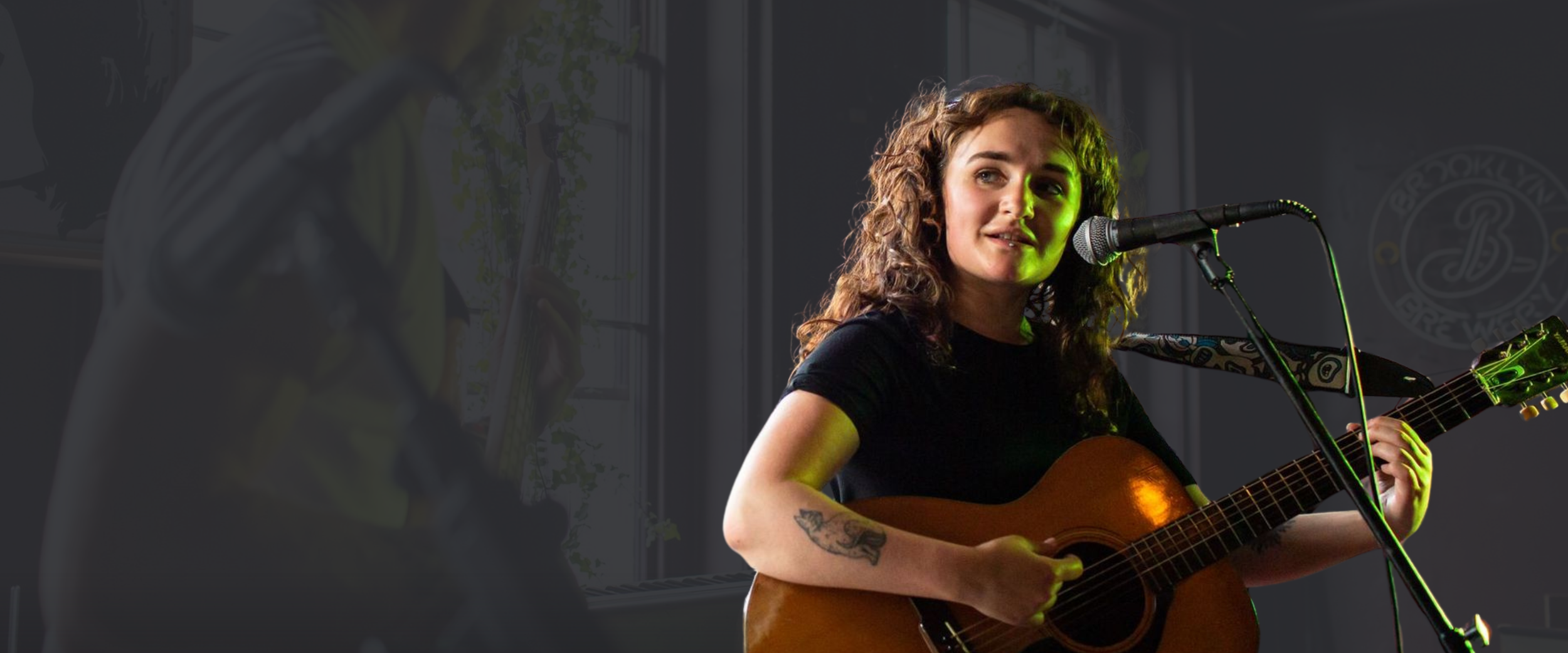 Young woman with curly hair playing acoustic guitar and singing into a microphone during a performance, with another musician partially visible on the left in an indoor venue.