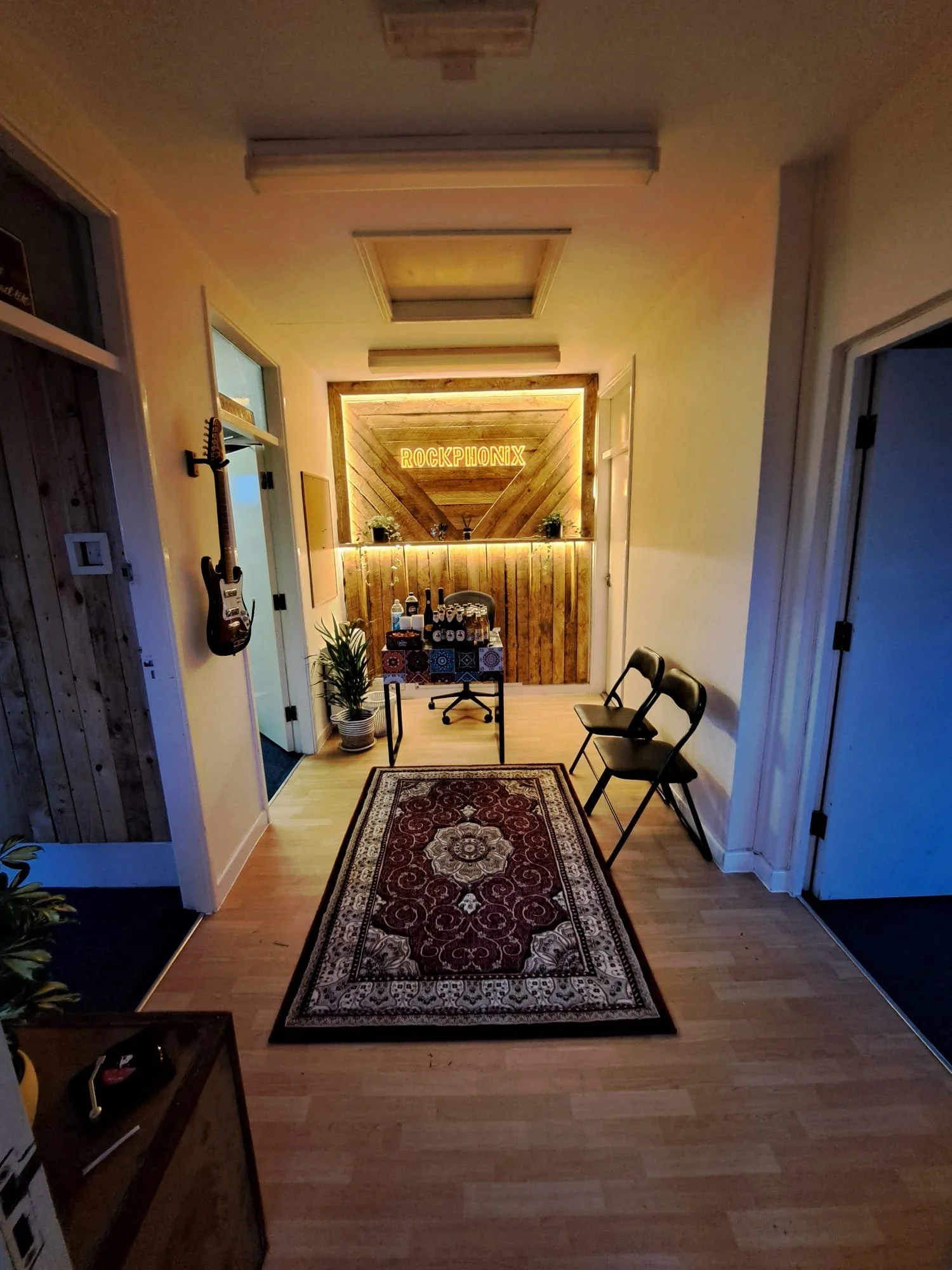 A small indoor space with a wooden decorated counter and a neon sign that says 'ROCKPHONIX'. There are three black folding chairs on the right, a patterned rug on a light wood floor, and a guitar hanging on the left wall. The space is cozy with potted plants and warm lighting.
