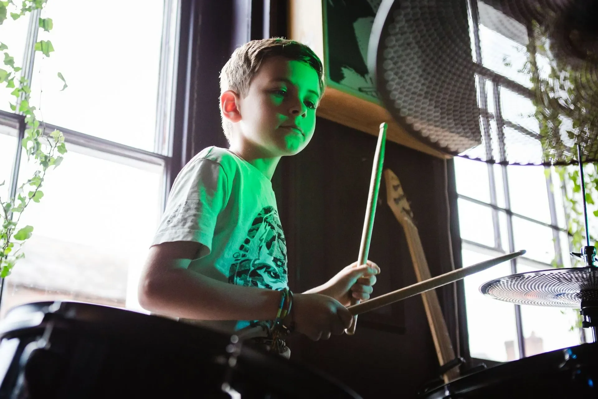 A young boy playing drums indoors, illuminated by natural light from a large window nearby.