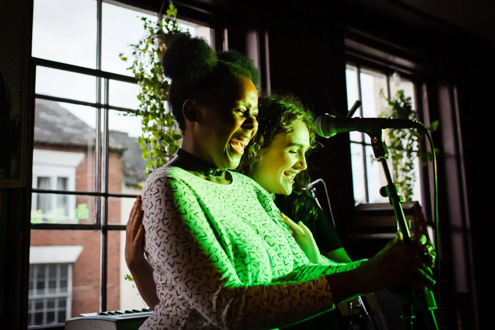 Two women smiling and singing into a microphone, with one holding a piano key, in front of large windows with natural light, and plants in the background.