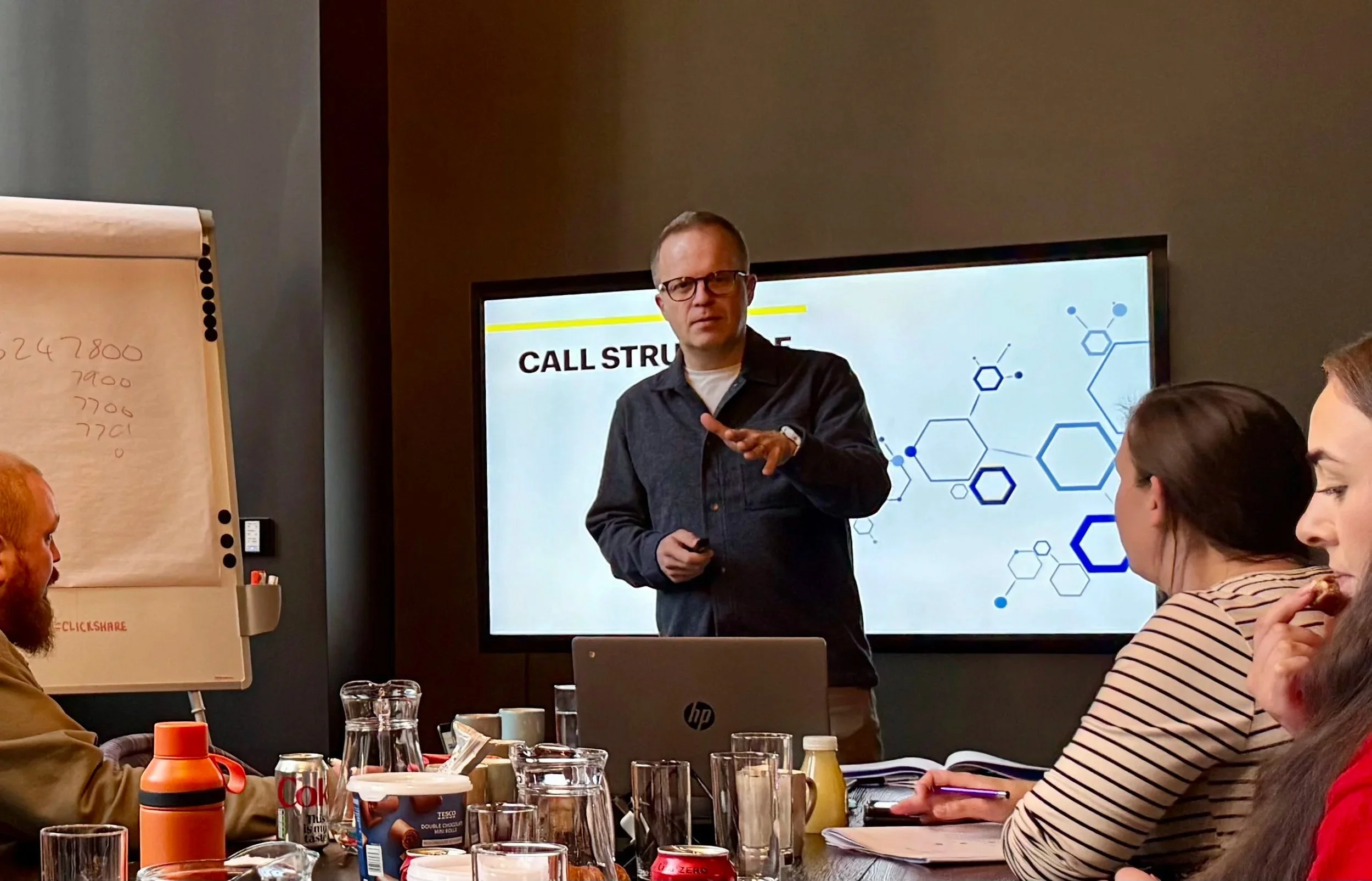 A man is giving a presentation on call structure in a conference room, with a screen behind him displaying a slide and several audience members listening and taking notes.