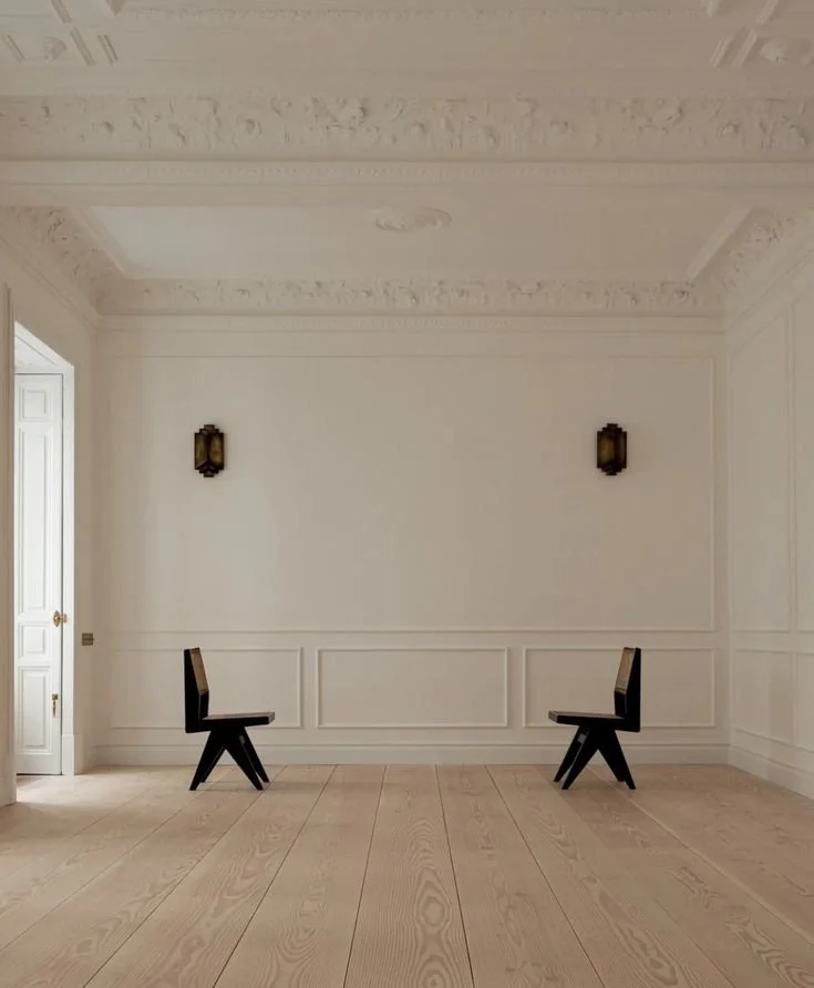 Salle élégante avec murs blanc, moulures ornementales, deux chaises noires modernes, et deux appliques murales en bronze. Sol en bois clair, porte blanche sur la gauche.