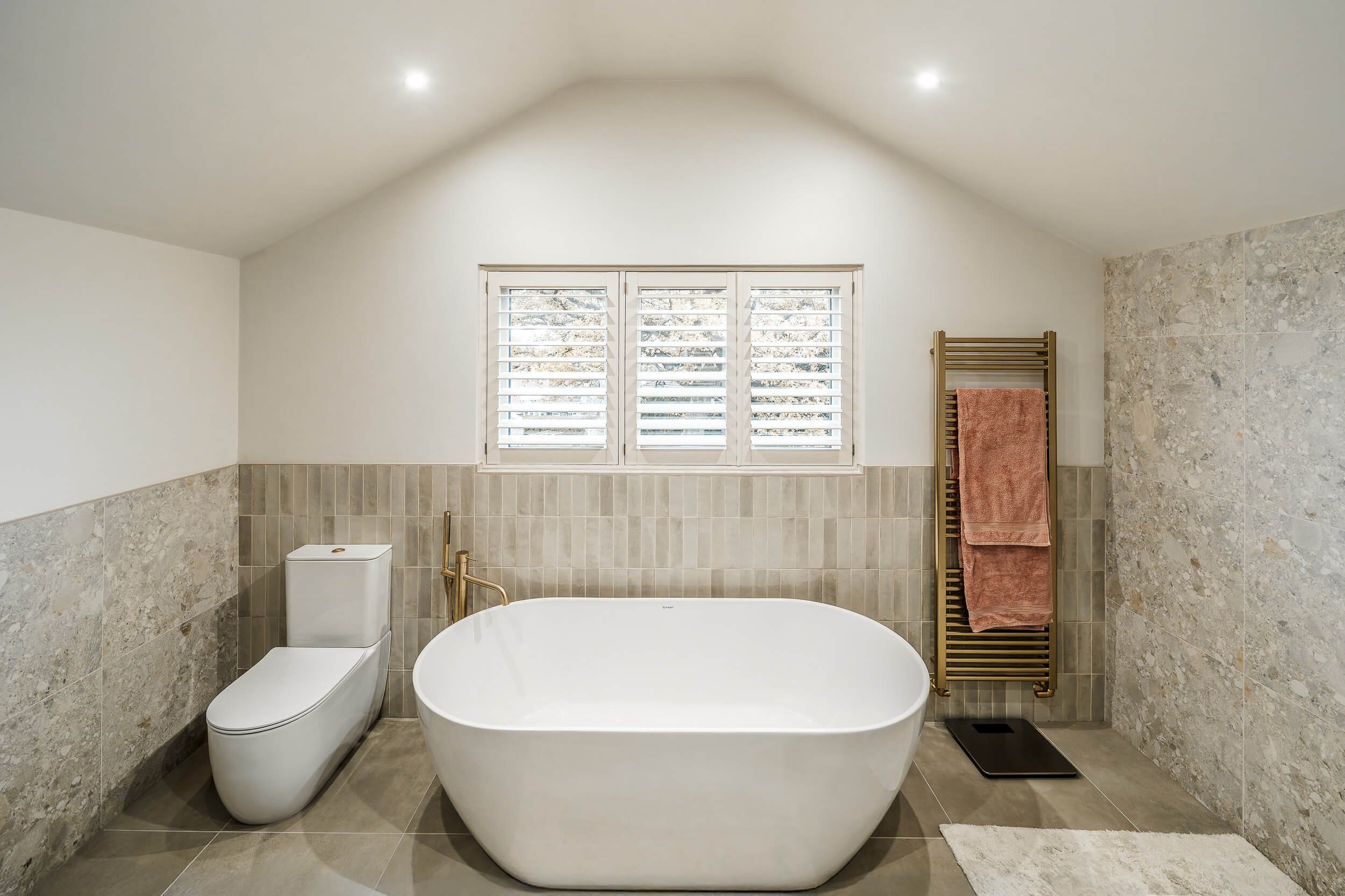 A modern bathroom with a freestanding bathtub, a toilet, and a tiled wall, with a window with shutters and a heated towel rack.