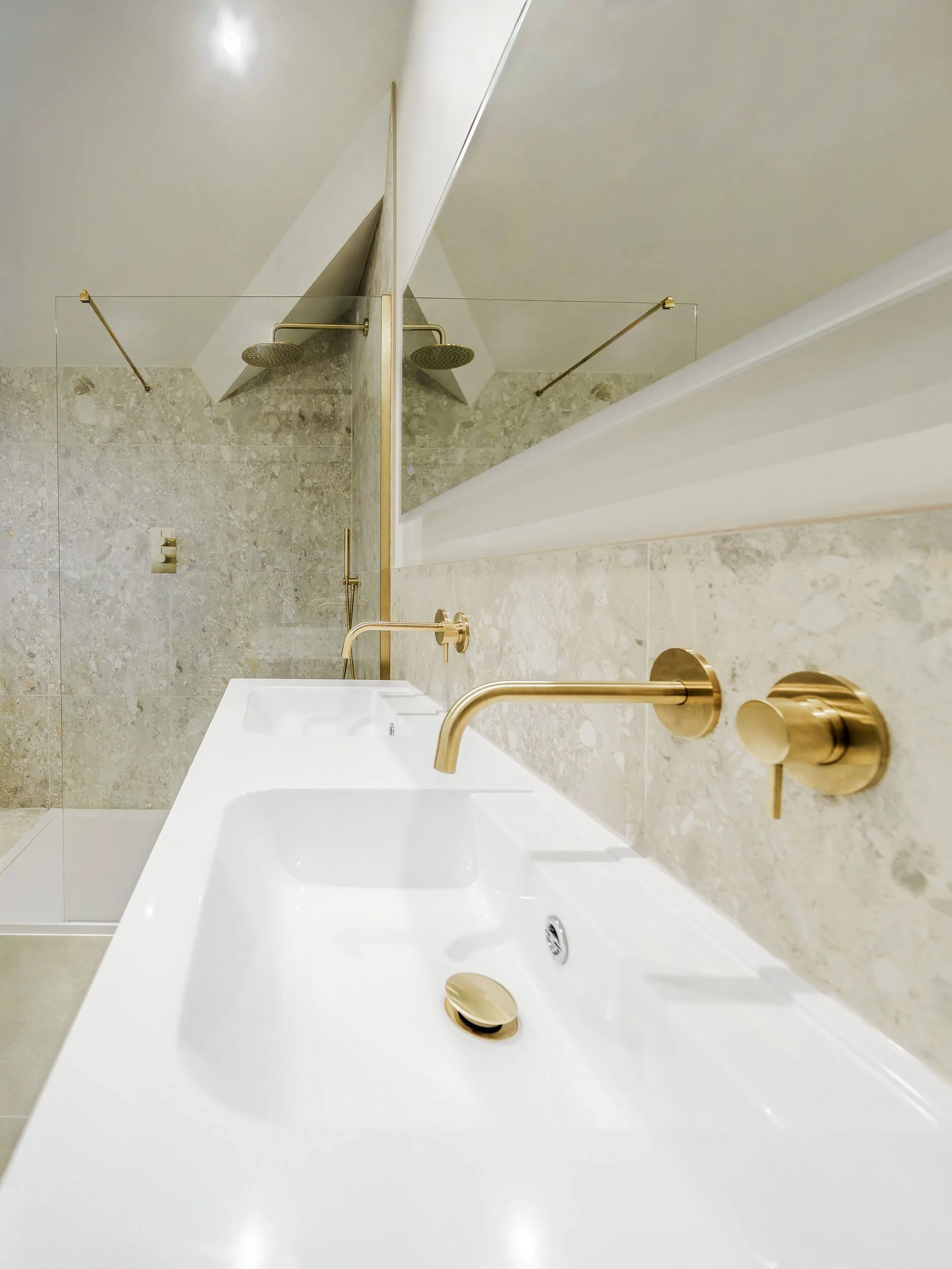 Modern bathroom with double sinks, gold fixtures, marble wall, and glass-enclosed shower with rain showerheads.