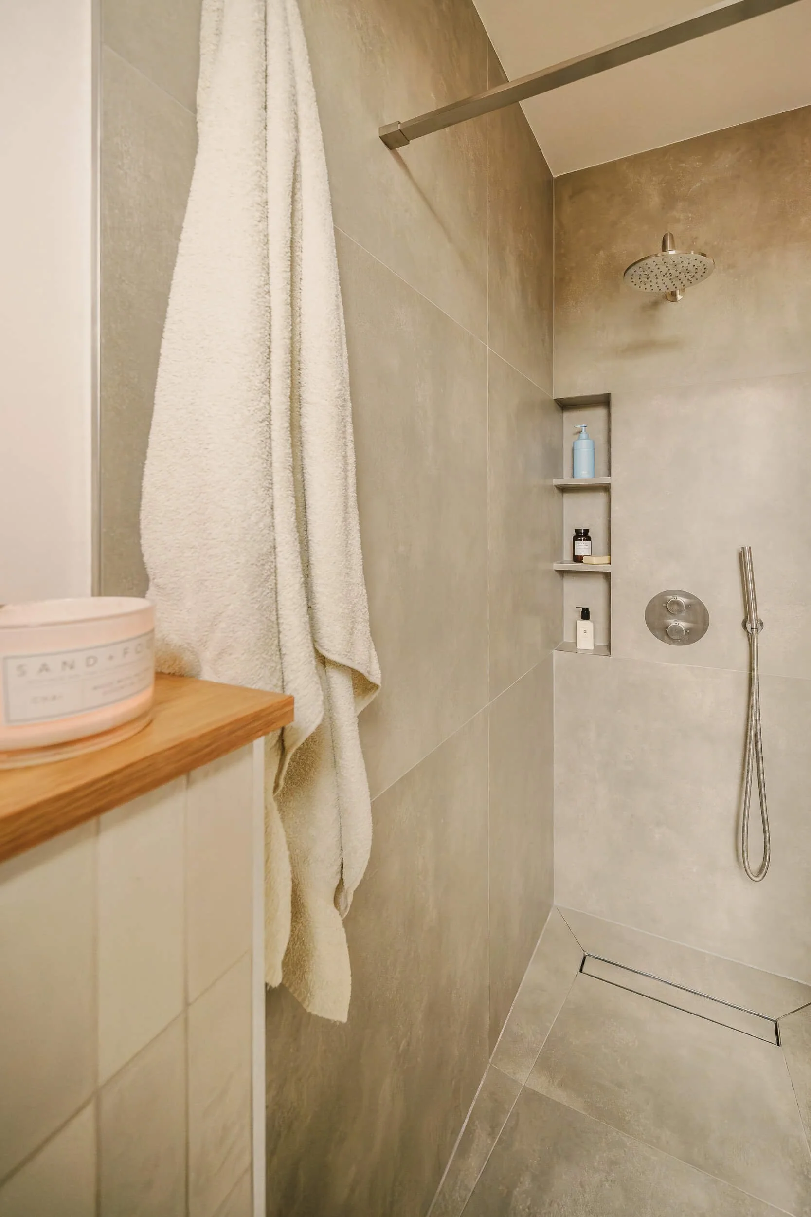 Modern shower with beige tiles, a rain showerhead, a wall-mounted handheld shower, built-in shelves with toiletries, and a white towel hanging.