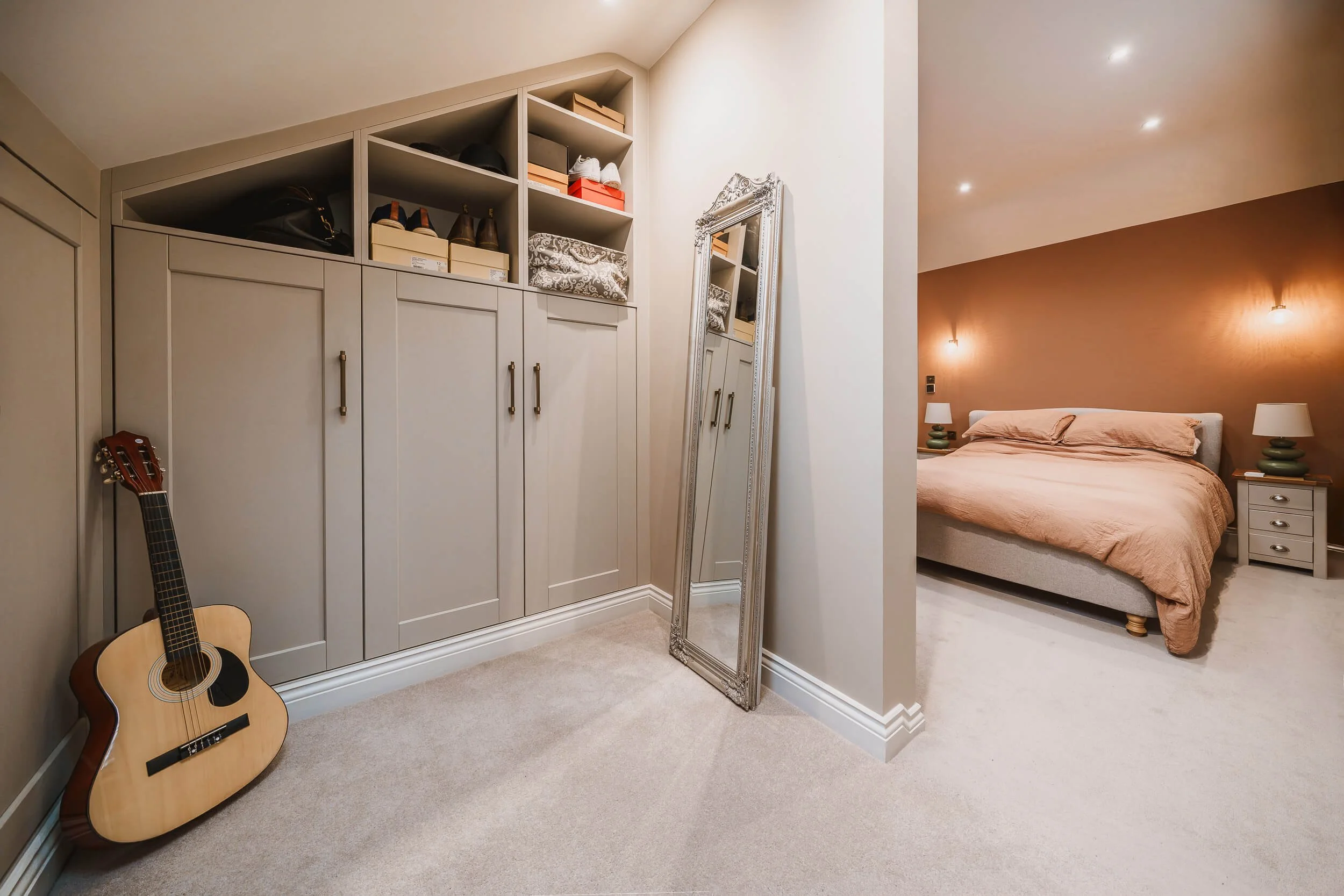 A bedroom with a beige bed, two nightstands with lamps, a large mirror leaning against the wall, built-in closet with boxes and shoes, and an acoustic guitar resting on the beige carpet.