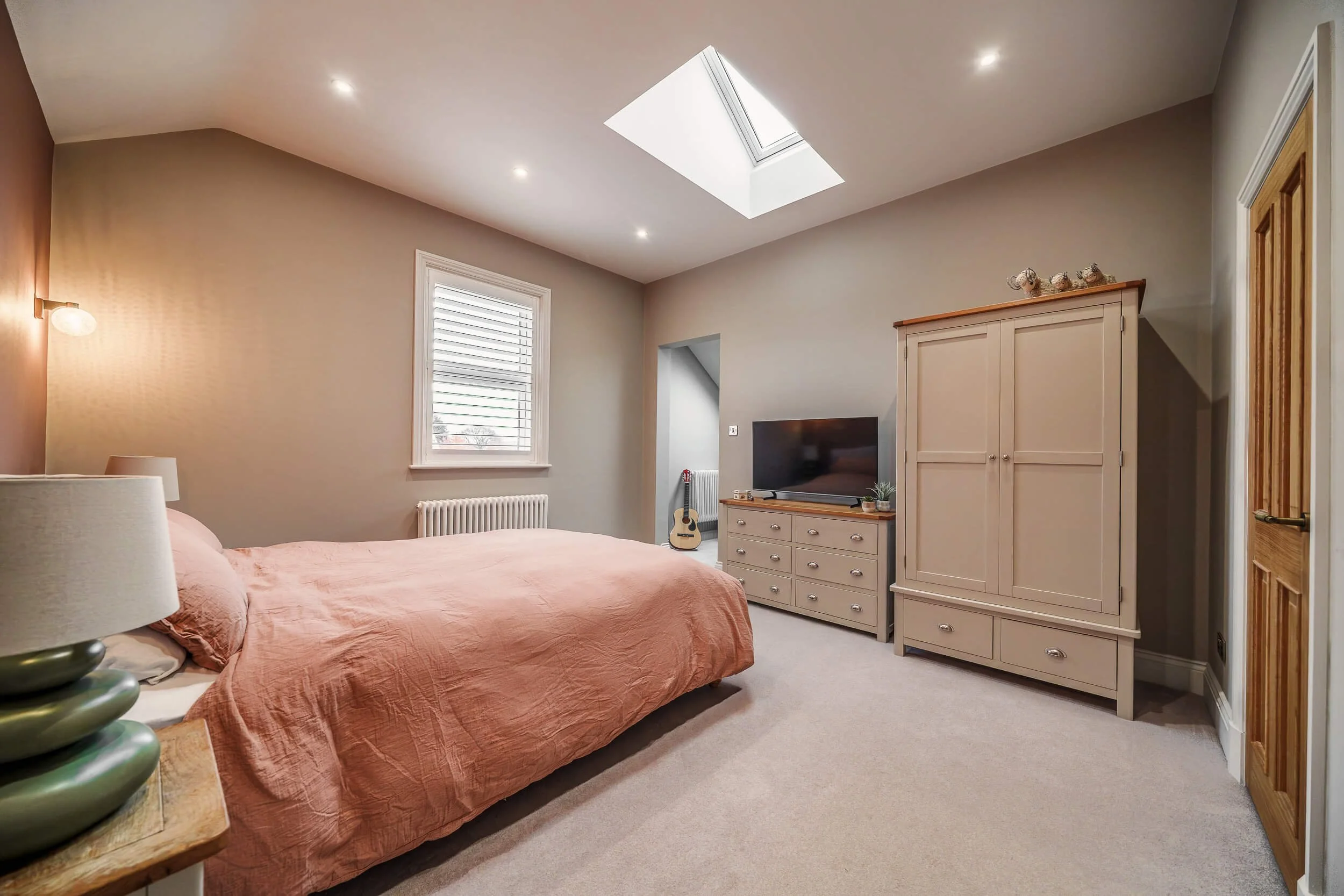 A bedroom with a pink bedspread, a window with white blinds, a ceiling skylight, a beige dresser with a TV, a guitar in the corner, a wardrobe, and a wooden door.