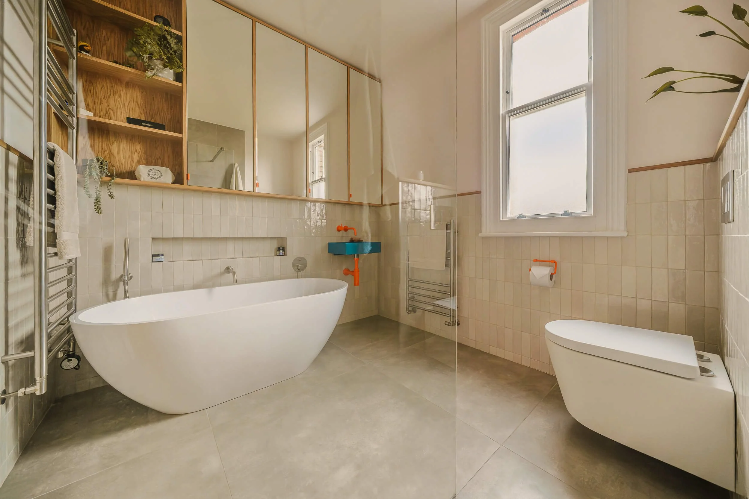 Modern bathroom with a white freestanding bathtub, a small blue sink with orange fixtures, a wall-mounted toilet with an orange toilet paper holder, a large window, and wooden shelving with decorative plants and towels.