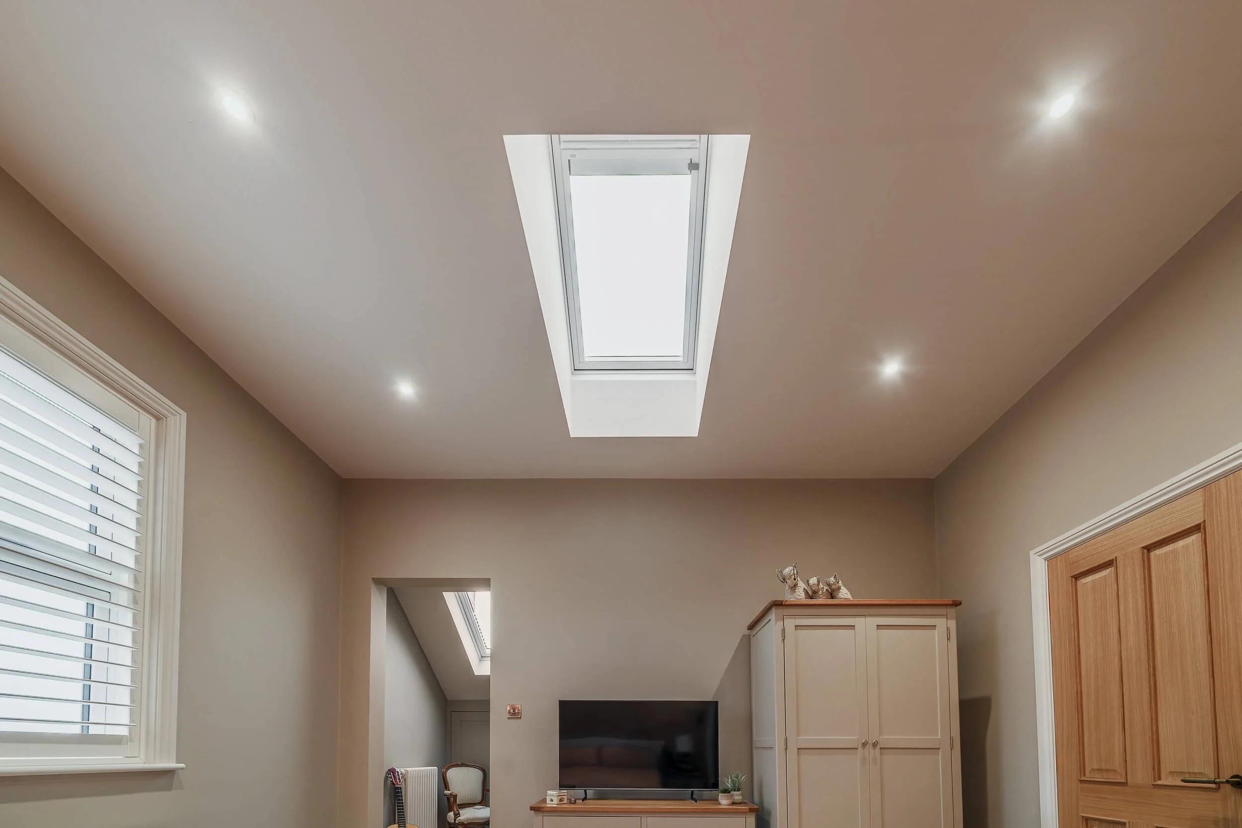 A room with a ceiling skylight, beige walls, a window with white blinds, a flat-screen TV on a wooden stand, a white wardrobe with decorative animal figurines on top, and a small patch of a doorway leading to another room with a chair.