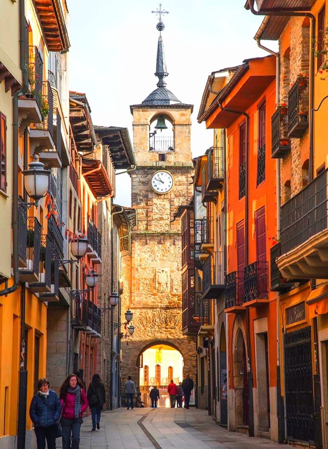 Vista de una calle peatonal en un pueblo con edificios coloridos y balcones de hierro forjado, en el fondo se encuentra una torre con campana y reloj, y un arco en la base de la torre.
