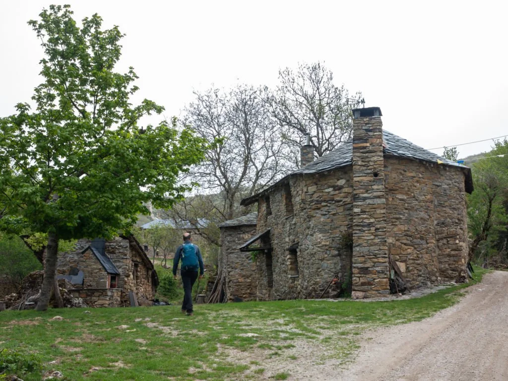 Persona caminando cerca de casas de piedra antiguas en un entorno rural con árboles y camino de tierra