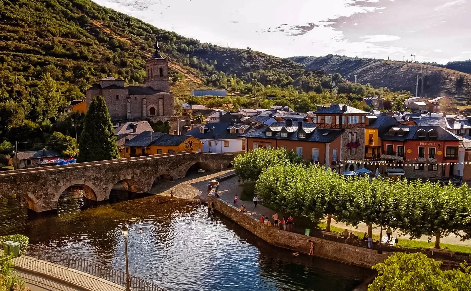Paisaje de un pueblo con un río, un puente de piedra, casas colorful y un fondo de colinas verdes bajo cielo nublado.